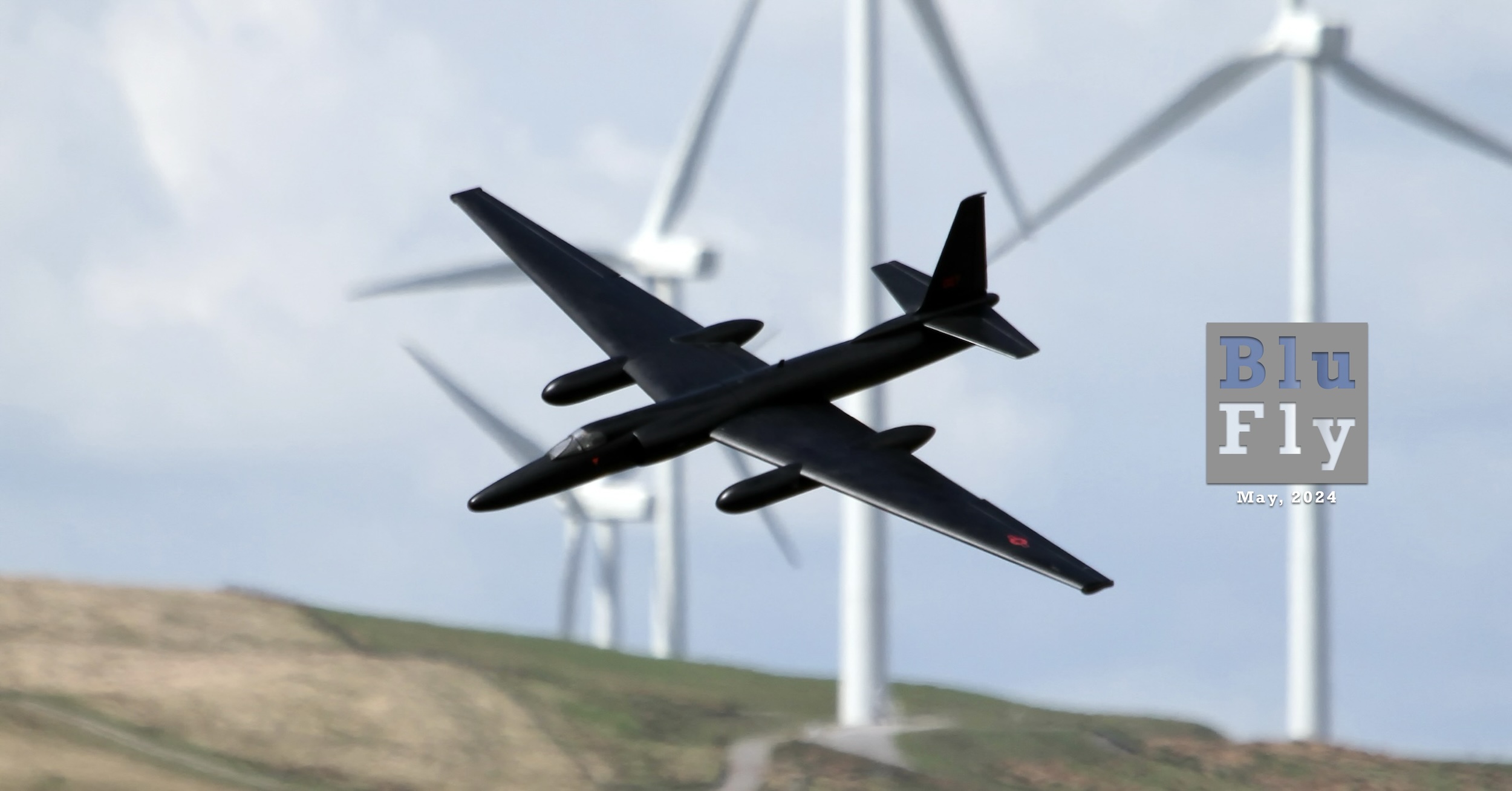 An all black radio-controlled model of the Lockheed U-2 spy plane is pictured flying in front of the wind farm located at Bwlch, South Wales, UK in May of 2018. The picture is credited to Phil Cooke. The sky is blue dotted with white clouds and the rolling hills are various shades of brown and green. At the right of the photo's subject there is a grey, BluFly logo consisting of the words « Blu » on one line and then « Fly » on the next line.  Both words are contained within a grey box. The former is shaded blue and the latter is shaded white. Below the logo are the words « May, 2024 » in crisp white letters.