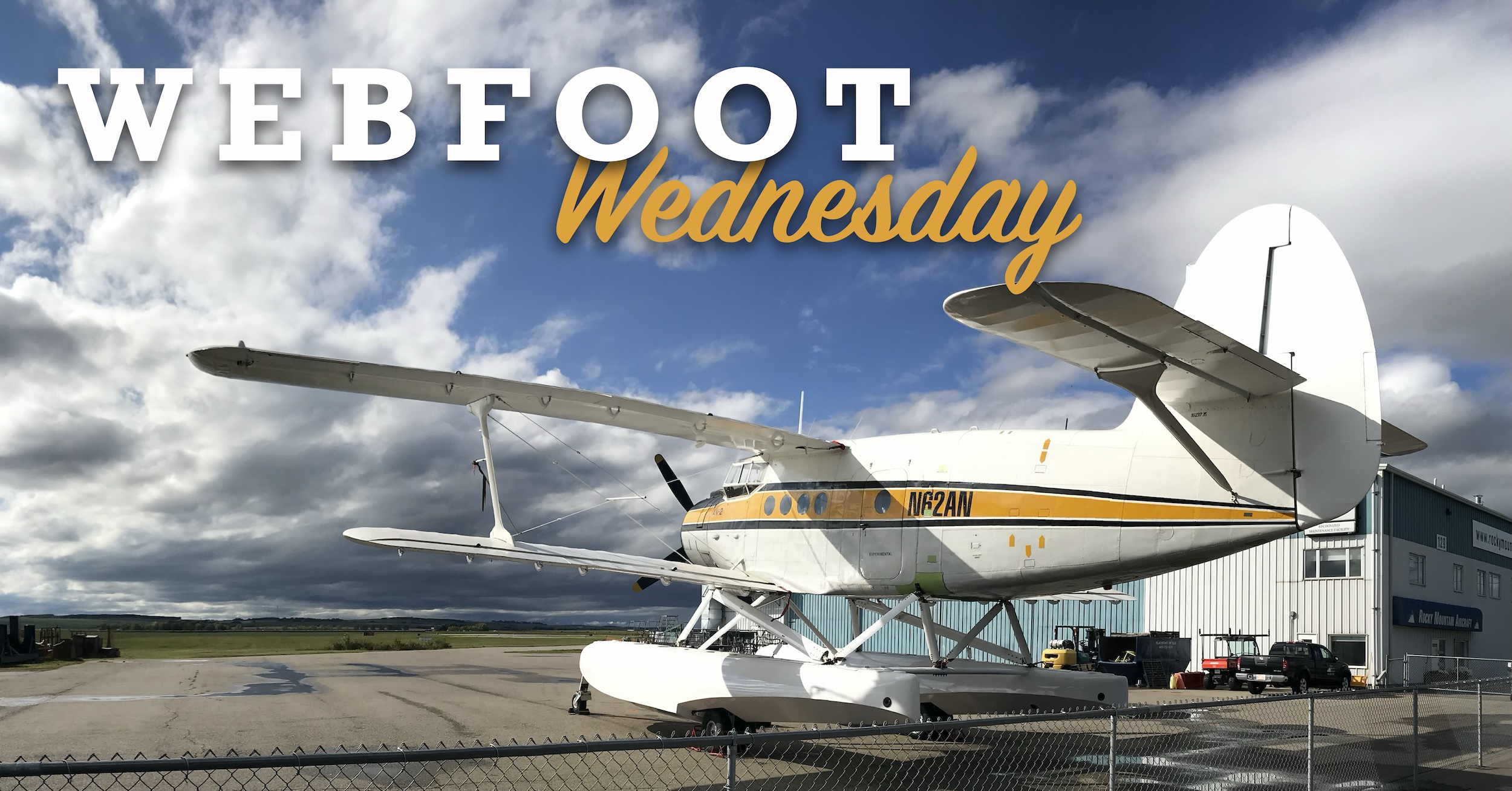 A large, white, fabric-covered biplane which is the subject of this post.  It is mostly white with a yellow and black accent stripes running the length of the fuselage.  The aircraft is mounted on two, large amphibious floats.  The sky in the picture is a vivid blue with fluffy white clouds.  The aircraft is parked in front of a hangar, which also has maintenance equipment parked in front of it. Superimposed over the image are the words « WEBFOOT » in large block letters in white and then the word « Wednesday » in script similar to the colour of the accent stripe of the plane.