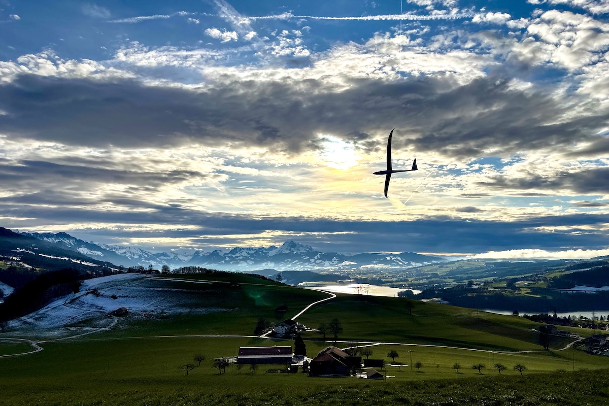 A radio-controlled glider flies in a stormy looking sky in Fribourg state, Switzerland. Below the aircraft is a rural, agricultural landscape. A lake is visible distance as are snow-covered mountains. (📸 Alexandre Mittaz)