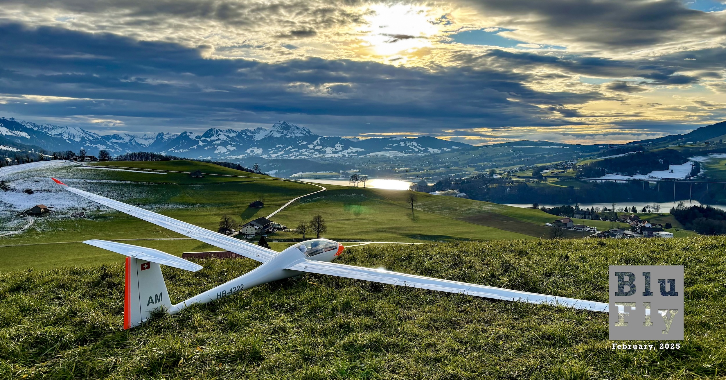 A radio-controlled model glider sits on a grass field. The camera position is behind, to the right and slightly above the glider. Its full 5.8 m span is captured within the frame. It is a modern glider seemingly built out of compostive materials. It has a narrow, high aspect ratio wing and T-tail. It is all white with bright red trim on the nose and on the trailing edge of the rudder. On the right side of the fuselage's tail boom the aircraft's registration « HB-4222 » is painted in black, block letters. The letters « AM » are painted in similarly styled letters on the vertical stabiliser. A Swiss flag is also painted on the vertical stabiliser. Appropriately scaled models of the pilots appear to have been placed in the cockpit. Beyond, in the middle distance, is a manicured farmer's field and farm house along with a winding road leading up to it. Beyond that is a lake off which the sun is reflecting. Beyond the lake are snow-covered mountains. Above, the story looking sky is partially broken overcast with a hazy sun. Superimposed on the bottom right corner of the photo there is a grey, BluFly logo consisting of the words « Blu » on one line and then « Fly » on the next line. Both words are contained within a grey box. The former is shaded blue and the latter is shaded white. Below the logo are the words « February, 2025 ». (📸 Alexandre Mittaz)