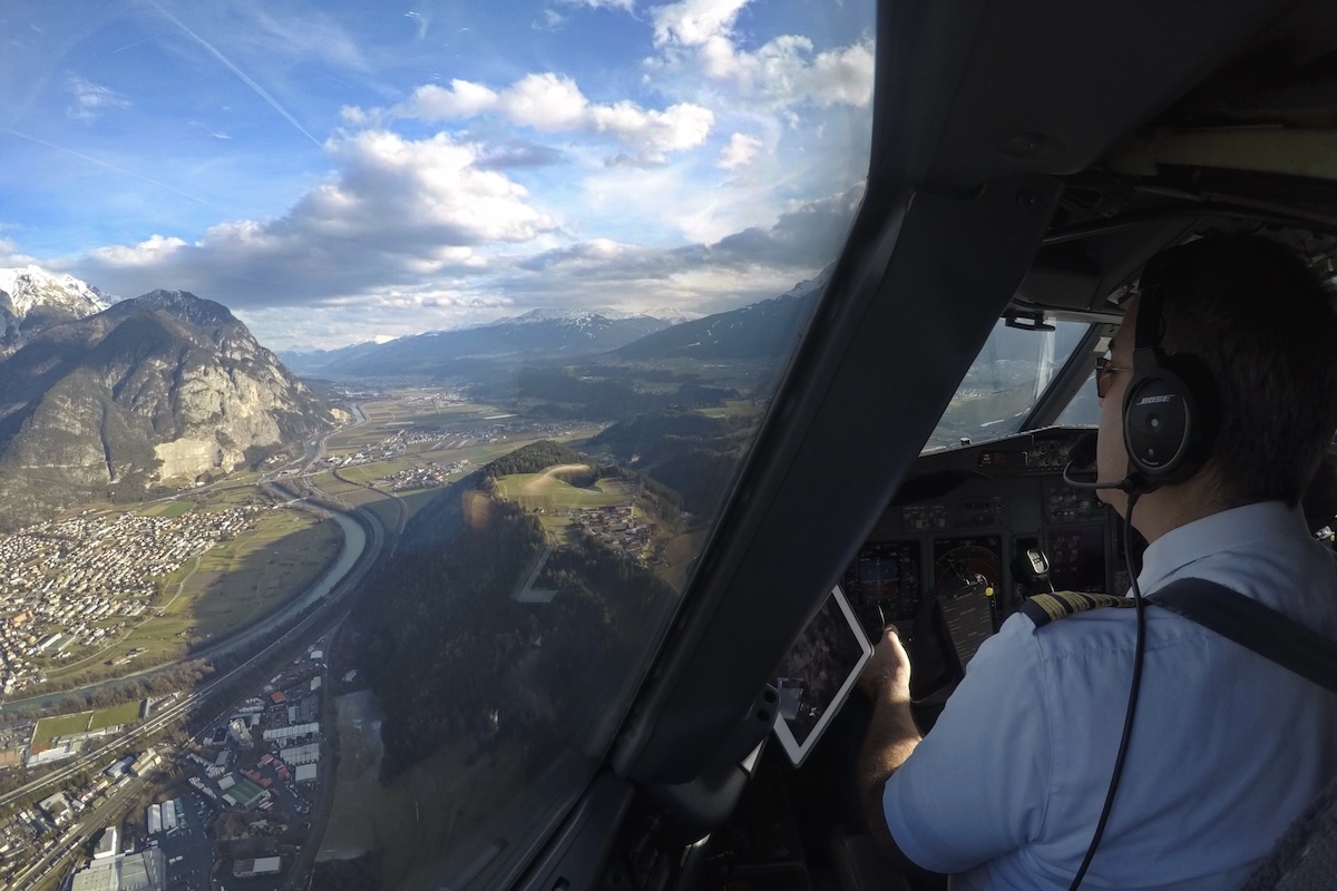 The interior of an airliner cockpit looking out the forward and port windows onto the approach to the airport at Innsbruck, Austria. The sky above is almost entirely blue with puffy white clouds at the horizon over the mountains. The pilot, seating in the left seat, is visible in the bottom right corner of the frame. (📸 Raymond Esveldt)
