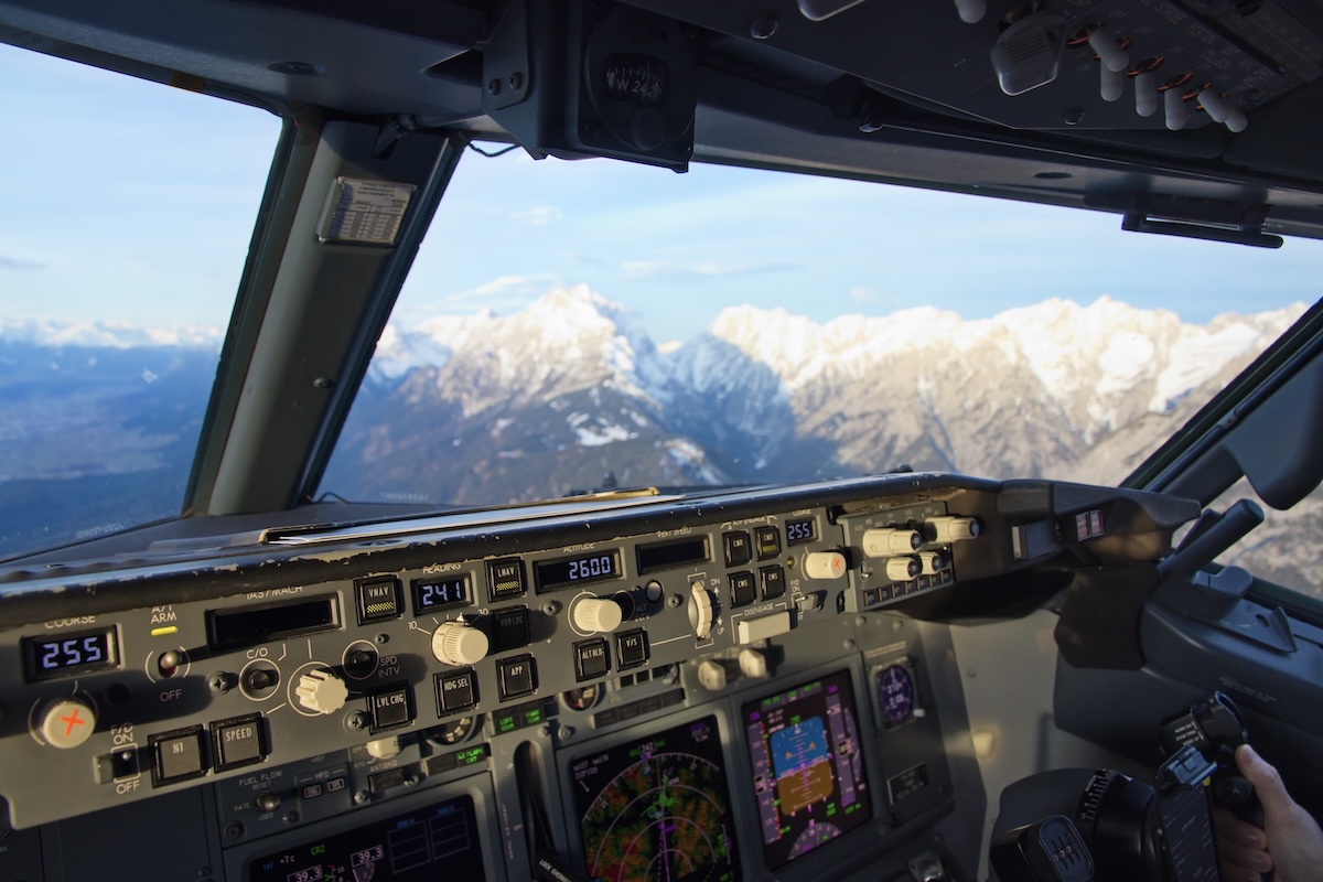 The interior of an airliner cockpit looking out the starboard windows onto the jagged, snow-covered mountains visible while on approach to the airport at Innsbruck, Austria. The sky above is almost entirely blue with layers of white clouds at the horizon over the mountains. A part of the aircraft's control panel is visible at the lower edge of the frame. (📸 Raymond Esveldt)