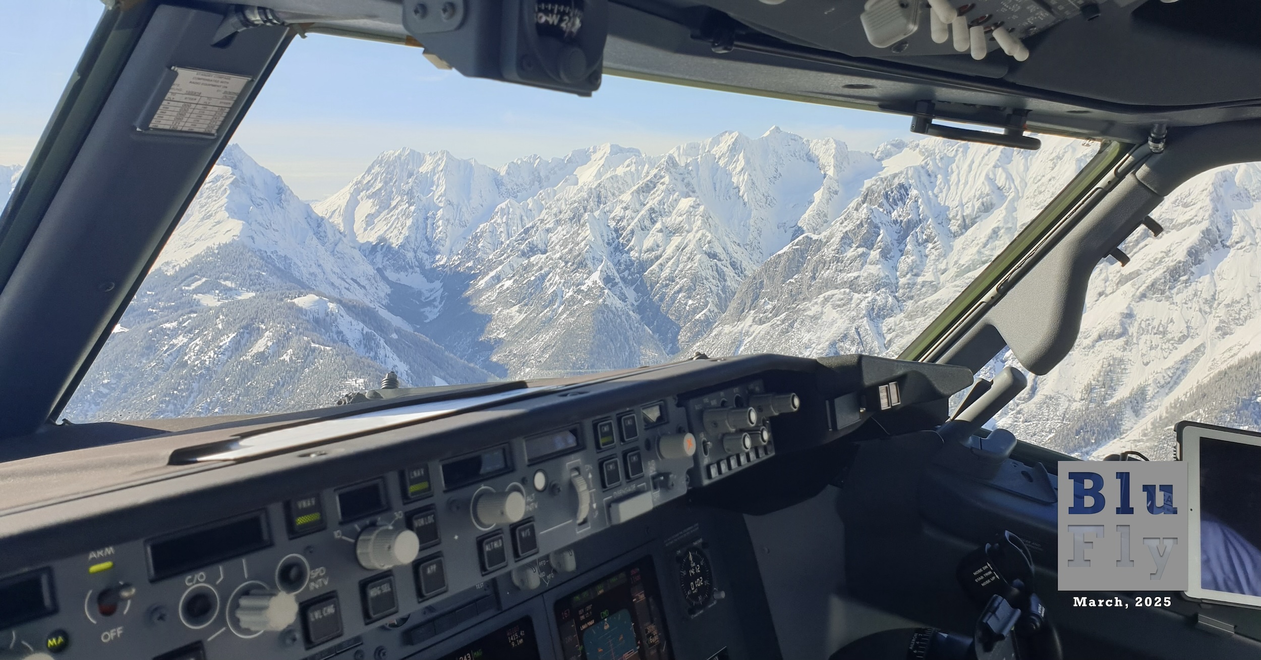The interior of an airliner cockpit looking out the starboard windows onto the jagged, snow-covered mountains visible while on approach to the airport at Innsbruck, Austria. The sky above is almost entirely blue with layers of white clouds at the horizon over the mountains. A part of the aircraft's control panel is visible at the lower edge of the frame. Original caption from source: « Flying into Innsbruck, Austria has a multitude of challenges. The peaks topping over 9,000 feet require pinpoint navigation accuracy to safely descend into the valley. The weather can be challenging with snow showers, icing conditions, low clouds over the city, or treacherous föhn wind creating severe turbulence and windshears. Engine-out performance is critical, requiring tailor-made engine-out departure routes through the winding valley. All aspects that make aviation interesting are combined at this one airport. But the amazing views make it all worth it! » (📸 Raymond Esveldt)