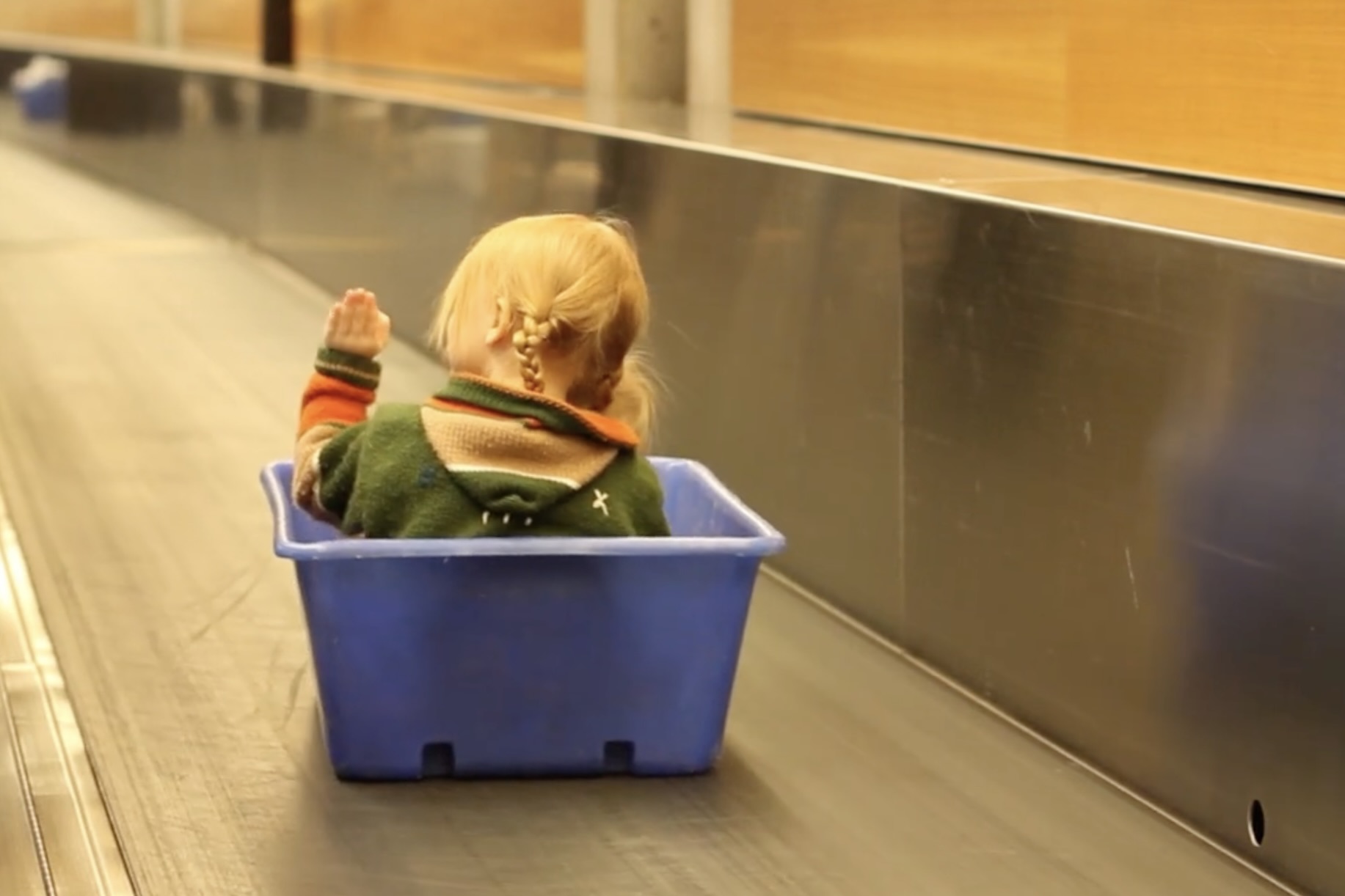 A small child rides a blue tote bin down a baggage belt as would be found at an airport. The child is waving to somebody not pictured in the frame. (📸 WestJet)