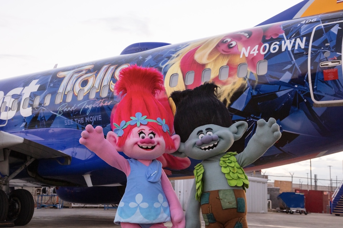 An image of the rear portion of a Southwest Airlines 737 fuselage which has been painted with a Trolls 'Band Together' motif from the DreamWorks animated film franchise. The registration number « N406WN » of the aircraft is painted in white block letters. In the foreground are two mascot-sized versions of the Poppy and Branch characters from the movie. (📸 Brianna Juda, Southwest Airlines)