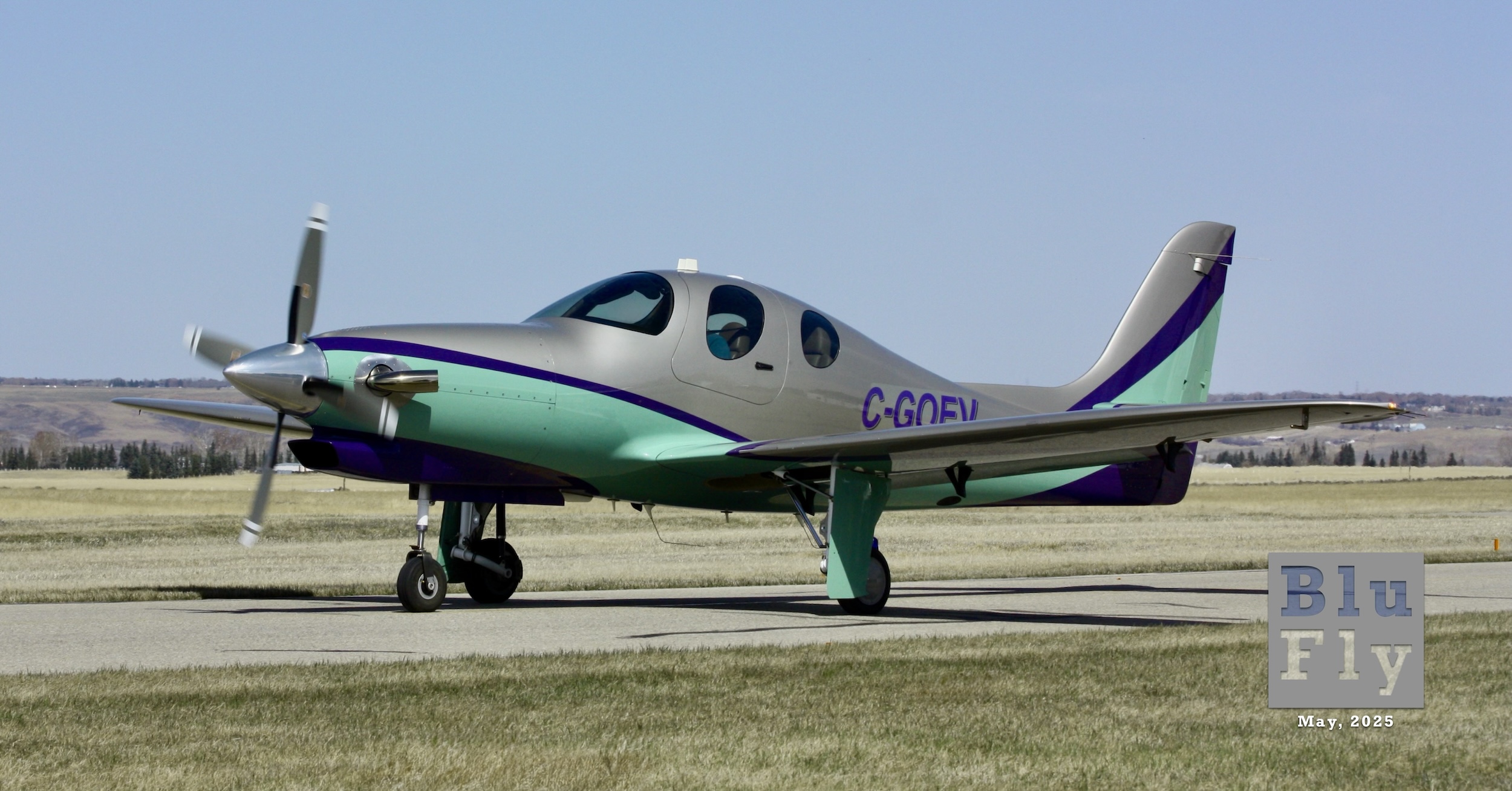 A Lancair Evolution taxiing from right to left along the taxiway just south of  Runway 08/26 at Springbank Airport near Calgary, Alberta, Canada on April 26, 2025. It is grey on the upper surfaces and minty green on the lower surfaces separated by a curvy dark blue or black stripe. The registration « C-GOEV » is clearly visible on the side of the fuselage. The weather appears to be sunny with clear stretching out to the horizon. In the distance, there are rolling hills dotted with small structures and patches of trees. Brown grass turning to green is visible both in front of and behind the taxiway. (📸 Terence C. Gannon)