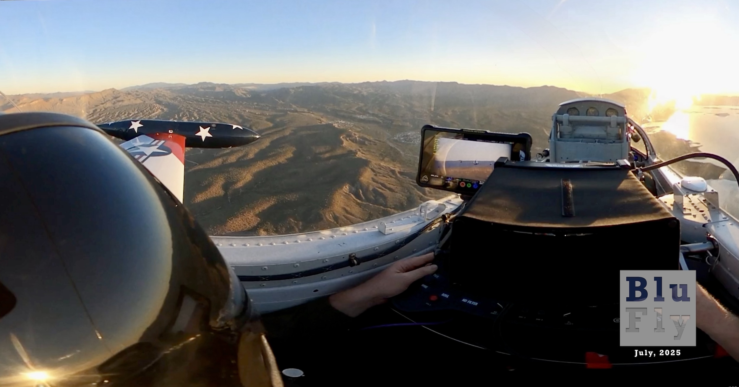 An image captured from the rear seat of a two-seat aircraft, with seats arranged fore and aft. At the left side of the frame is the helmet of the person sitting in the rear seat. They are reaching forward with their hands to equipment used to control the feature film cameras the aircraft carries. Forward of this position, it's possible to see the headrest of the person sitting in the forward pilot's seat, but not the actual pilot. The camera is positioned just to the right of the after seat's occupant's head. It's a wide-angle shot with views out both the right and left sides of the bubble canopy. The predominant view is out the left, which reveals a rolling, desert hills landscape. The tip tank at the end of a straight wing is also visible. The tip tank is painted dark blue with white stars. The wing is painted red and white with a US Air Force roundel partially visible. Out the right side, a sunlit body of water can be seen. The sky is completely clear, and the sun is on the horizon on the right side of the frame. A smallvideo screen is visible between two seats, and close examination reveals two or three aircraft being captured in the frame, presumably with the cameras mounted on this aircraft. Superimposed on the bottom right corner of the photo is a grey, BluFly logo consisting of the words « Blu » on one line and then « Fly » on the next line. Both words are contained within a grey box. The former is shaded blue, and the latter is shaded white. The logo letters are ‘see-through' such that elements of the photo can be seen through them. Below the logo are the words « July, 2025 » in a similar typeface in white. The caption from the source reads: « Our glorious cover photo this month is from the rear seat of Film Knight 2, a Lockheed T-33 lovingly known as 'Stars and Stripes'. Up there, behind the forward headrest, is Pilot-in-Command Robert 'Scratch' Mitchell, and in the rear seat controlling the cameras is Director of Photography Peter Degerfeldt. It was captured in April 2024 near Lake Roosevelt, Arizona. » (📸 Robert 'Scratch' Mitchell | The Film Knight)