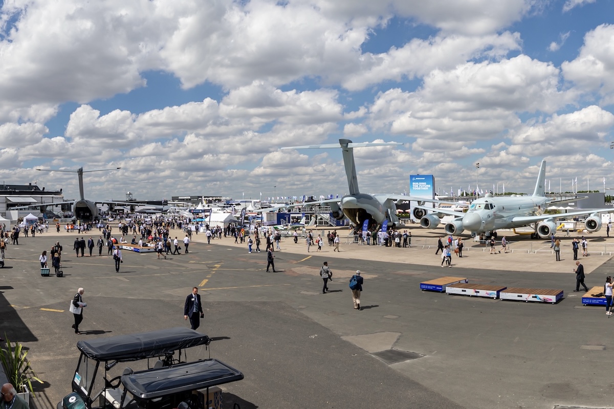 An image of one of the main static exhibit areas at the Paris Air Show in 2019. It captures a wide variety of both military and civil aircraft along with many people milling around them. The sky above is vivid blue with many cumulus clouds. From the original source: « This image is a panorama consisting of multiple frames that were merged or stitched in software. As a result, this image necessarily underwent some form of digital manipulation. » (Matti Blume via Wikimedia under CC BY-SA 4.0, cropped)