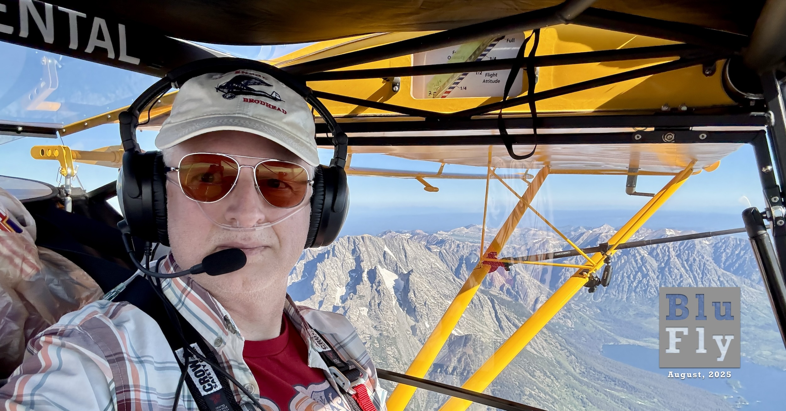 A man sits at the controls of a small, yellow aircraft while it is in flight. He is turned to the right, slightly, so the picture captures him, the interior of his aircraft as well as the view out the left side of the aircraft. The latter is of a mountain range stretching out to just below the horizon. The horizon itself is somewhat blurred. The sky above is gin-clear blue. Superimposed on the bottom right corner of the photo is a grey, BluFly logo consisting of the words « Blu » on one line and then « Fly » on the next line. Both words are contained within a grey box. The former is shaded blue, and the latter is shaded white. The logo letters are ‘see-through' such that elements of the photo can be seen through them. Below the logo are the words « August, 2025 » in a similar typeface in white. Original caption from source: « Selfie flying the plane. I have an oxygen cannula in my nose, sunglasses, and headphones on. The North Tetons and Jenny Lake are in the background. » (📸 Dan Yocum)
