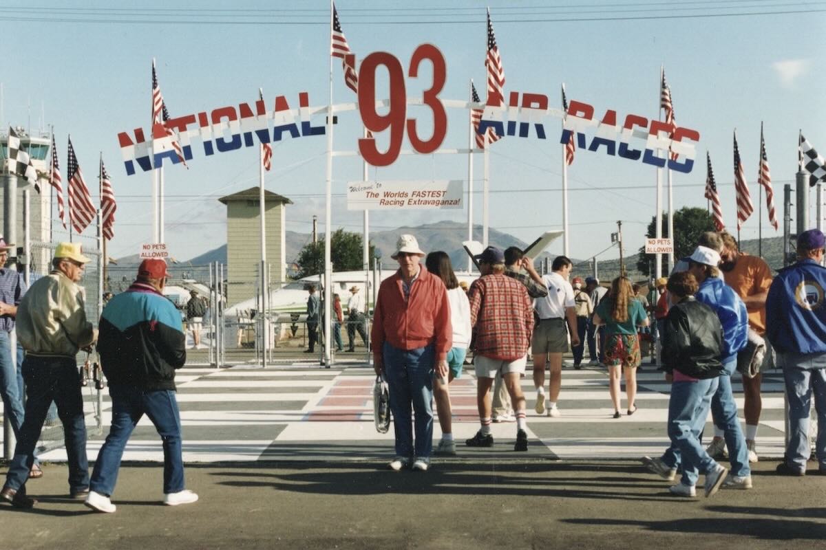An unsmiling Patrick Gannon, in an orange jacket, is pictured at the entrance to the 1993 Reno Air Races held at Reno-Stead Airport. He is wearing a beige bucket hat and is looking directly at the camera. There is an arch over the walkway that reads « NATIONAL '93 AIR RACES » in large block letters painted in a red, white, and blue motif. Below that is a much smaller sign that says « Welcome to the World's Fastest Racing Extravaganza ». On either side of the arch are multiple United States and chequered flags. Behind him, there are other attendees at the event, and beyond them, aircraft are also pictured. It is a sunny day with a clear blue sky with a few white, puffy clouds. Based on how people are dressed, it's fairly warm.