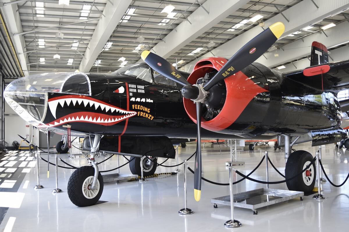 A pristine, black A-26 'Marauder' aircraft is pictured in a well-lit indoor display space. It is oriented so that it is pointing to the left side of the frame and slightly toward the camera position. It has a stylised shark's mouth and eyes (like a P-40) as well as the words « FEEDING FRENZY » painted on the nose just aft of a clear enclosure making up the nose. The aircraft has red accents painted on the leading edge of the engine nacelles as well as the top of the vertical stabiliser. Original caption from source: « She was then sold to Gower Lebel Inc in Seattle and was repainted in a black USAF colour scheme before moving to California in May 1990 with new owners A&T Productions. She was reregistered as N34538 that September, fitted with a glazed nose from an A-26C and given her current name “Feeding Frenzy”. She was sold to an owner in Kelowna, British Columbia, in March 1997 before joining the museum here in April 1999. ¶ Lyon Air Museum, John Wayne Airport, Orange County, California, USA.¶ 16th March 2018 » (📸 Alan Wilson via Flickr under CC BY-SA 2.0)