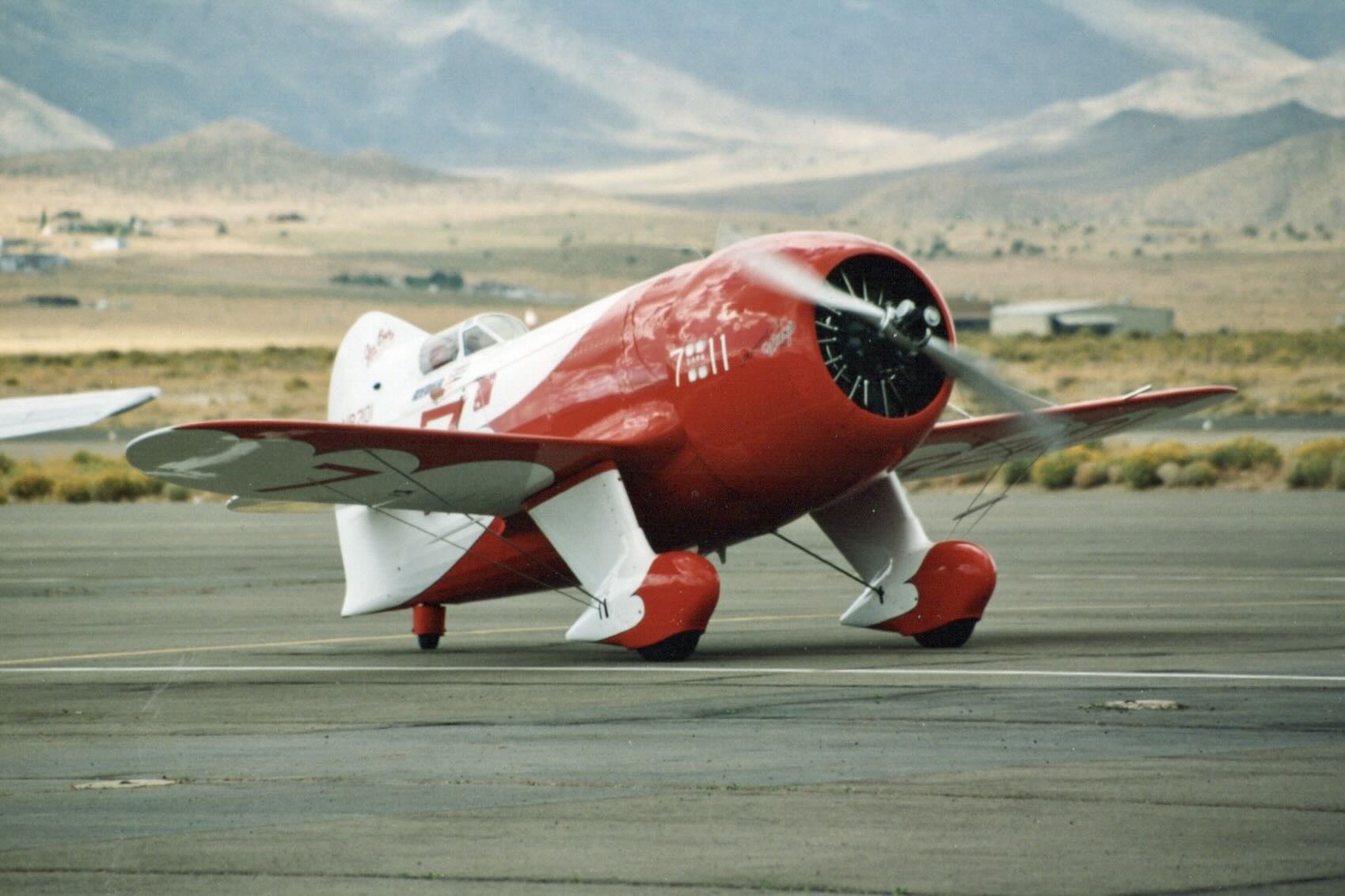Delmar Benjamin's read and white Gee Bee R-2 is pictured either on a runway or taxiway at Reno-Stead Airport at the Reno Air Races in 1993. The aircraft is pointing to the right and slightly towards the camera position. It's just possible to see the top of the pilot's head through the canopy. The engine is running. Rolling, desert hills are visible in the distance as well as some small airport structures.