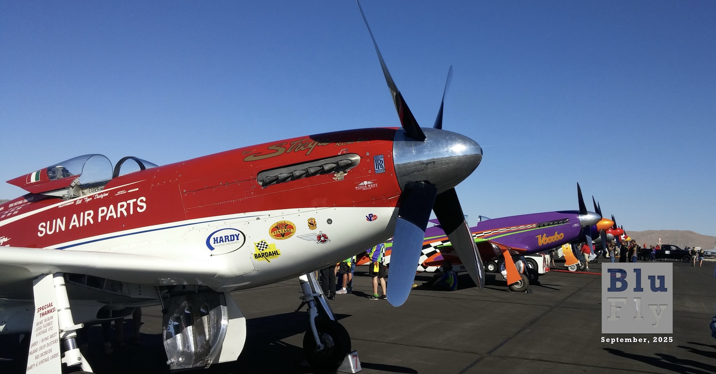 A colourful line-up of P-51 'Mustang' aircraft modified for air racing. None of the visible liveries are military in nature but rather various attention-grabbing civilian colour schemes. They are pointed to the right side of the frame. The aircraft nearest the camera, which is red on the upper surfaces of the fuselage and white on the lower surfaces and white wings, has the word « Strega » painted above the exhaust stacks in gold, stylised script. The words « SUN AIR PARTS » are painted below the cockpit. The aircraft beyond this is painted mostly purple and has the word « Voodoo » painted in orange-y letters below the exhaust stacks. Both aircraft have chrome spinners. It's not possible to see any further, significant detail beyond these two aircraft. The sky above is gin-clear blue with bright sun. The tarmac on which the aircraft are parked is dark grey and smooth. A few service vehicles and people are also visible. Original caption from source: « Reno 2015 Unlimited Gold Line Up, Strega, Voodoo, Rare Bear, Dreadnaught » (📸 Reno 2015 Unlimited Gold Line Up by D Ramey Logan.jpg from Wikimedia Commons by Don Ramey Logan, CC-BY 4.0) »