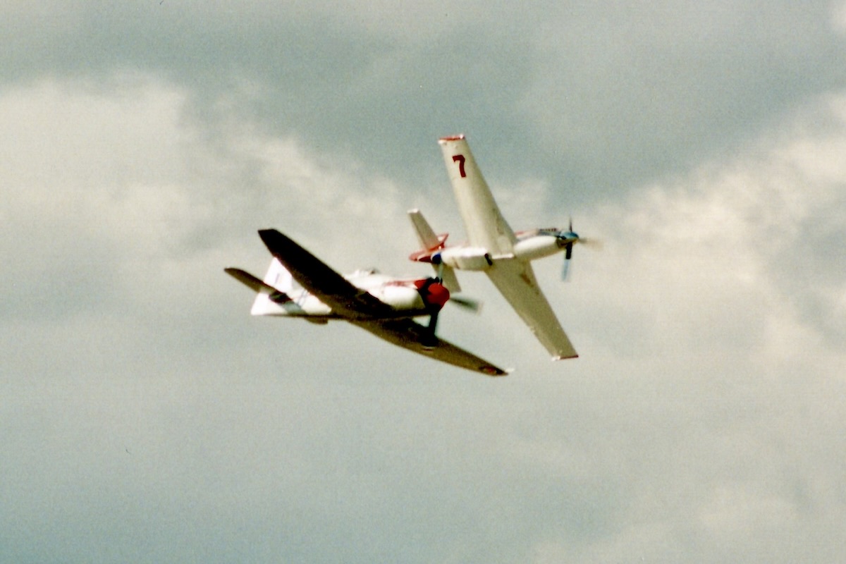 Two aircraft fly in close proximity to each other. They are framed against a cloudy sky with no blue visible. The first of the two aircraft is Bill Destefani's 'Strega', a modified P-51 'Mustang' otherwise known as Race 7. Below, and slightly to the left of this is Dennis Sanders' 'Dreadnaught', a modified Hawker 'Sea Fury' otherwise known as Race 8. The aircraft are racing from left to right in the frame.