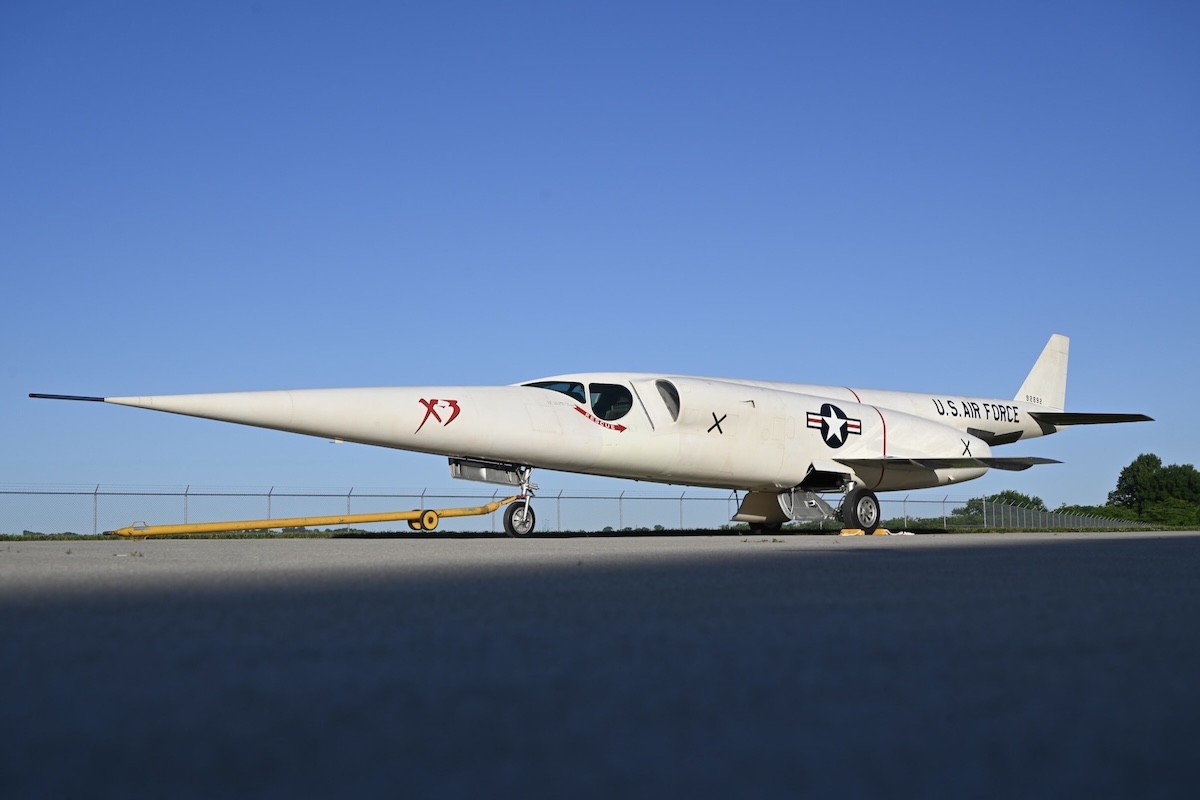 A needle-sharp, white, X-3 'Stiletto' aircraft is parked on grey tarmac under a gin-clear blue sky. The letters « X-3 » are painted in stylise red type on its nose. It also has a US Air Force roundel painted just about its short, stubby wings. The words « US Air Force » are painted on the tail boom. A barbed-wire fenced is visible in the background and a group of trees is visible on the right. Original caption from source: « DAYTON, Ohio -- Douglas X-3 Stiletto resting outside of the R&D Gallery at the National Museum of the USAF on May 28, 2024. » (📸 Ken LaRock, US Air Force via National Museum of the United States Air Force website in the public domain)