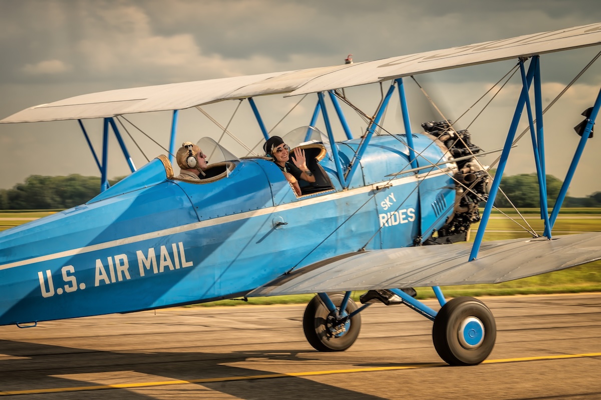 NC9079, a Travel Air E-4000 with a blue fuselage with either gold or brown wings, taxis from left to right and slightly away as it passes the camera position. The words « U.S. AIR MAIL » are painted in white on the side of the fuselage after the cockpit. The words « SKY RIDES » are painted on the side of the fuselage ahead of the cockpit. The pilot, in the rear seat, is looking forward over the engine. The passenger, in the front seat, is turning toward the camera and is both smiling and waving. There is green grass just beyond the taxiway. A line of trees is visible in the distance. The visible sky appears mostly cloudy but the sun seems to be shining. These aircraft were photographed at the 'Barnstormer Carnival' in Springfield, Ohio in July 2024. (📸 Richard 'Zulu' Souza | Full Disc Aviation)