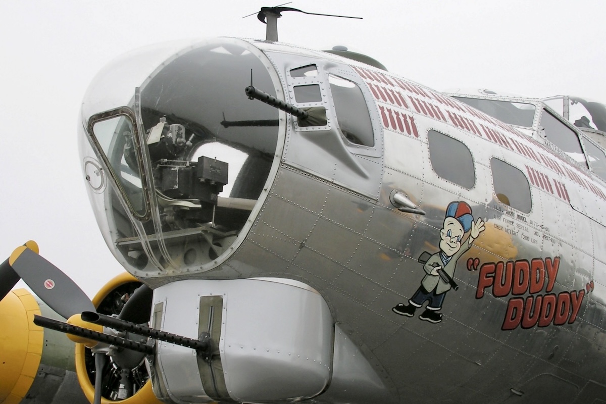 The nose of a restored B-17 aircraft is parked on an airport ramp. It is pointing toward and slightly to the left of the camera position. Painted on the left side of the fuselage is a character that looks like Elmer Fudd. Beside this are the words « "FUDDY DUDDY" ». Also painted on the side of the fuselage are the silhouettes of bombs, which seemingly indicate how many sorties this aircraft was on. (📸 Greg Goebel via Wikimedia Commons under CC BY-SA 2.0)