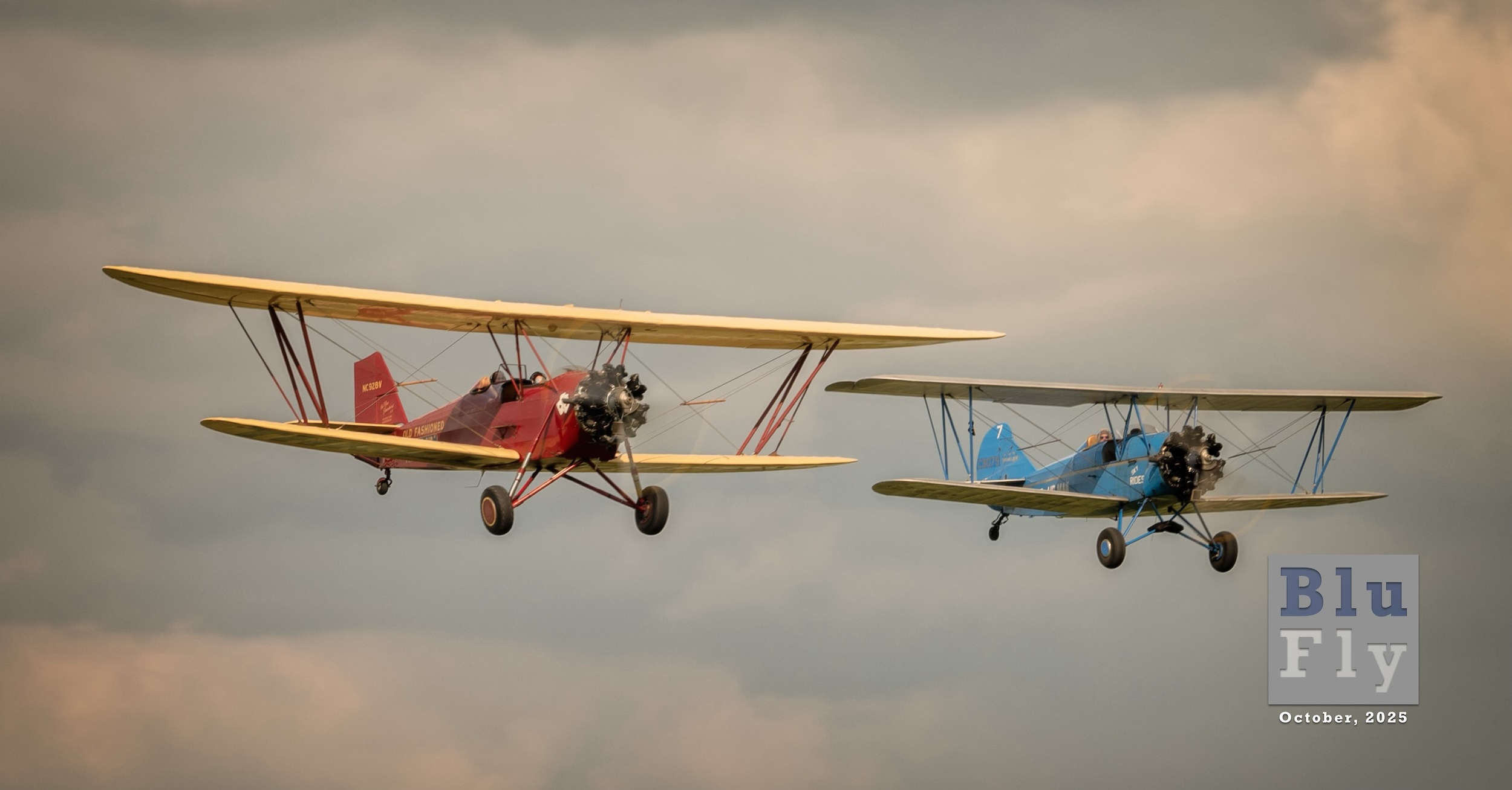 Two antique biplanes fly toward and to the right of the camera position set against a background of cumulus clouds. On the left is NC928V, a New Standard D-25 with a red fuselage and yellow wings. On the right is NC9079, a Travel Air E-4000 with a blue fuselage with either gold or brown wings. These aircraft were photographed at the 'Barnstormer Carnival' in Springfield, Ohio in July 2024. Superimposed on the bottom right corner of the photo is a grey, BluFly logo consisting of the words « Blu » on one line and then « Fly » on the next line. Both words are contained within a grey box. The former is shaded blue, and the latter is shaded white. The logo letters are ‘see-through' such that elements of the photo can be seen through them. Below the logo are the words « October 2025 » in a similar typeface in white. (📸 Richard 'Zulu' Souza | Full Disc Aviation)