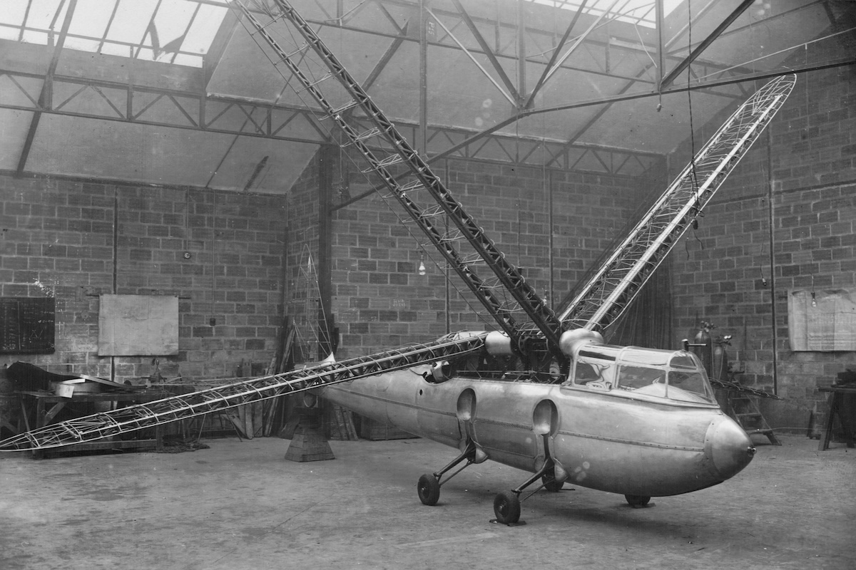 An old, black-and-white image of an Riout 102T 'Alérion'. It has a helicopter-like fuselage with the tail of a traditional aircraft. It has four thin wings which are flapped, as would be the case for a bird or insect like a dragonfly. In this picture they are bare framework, prior to being covered in some sort of material. The aicraft seems to be located in a construction shed with the framing of the building clearly visible along with other equipment found in such a locale. (📸 René Riout via Wikimedia Commons in the public domain)