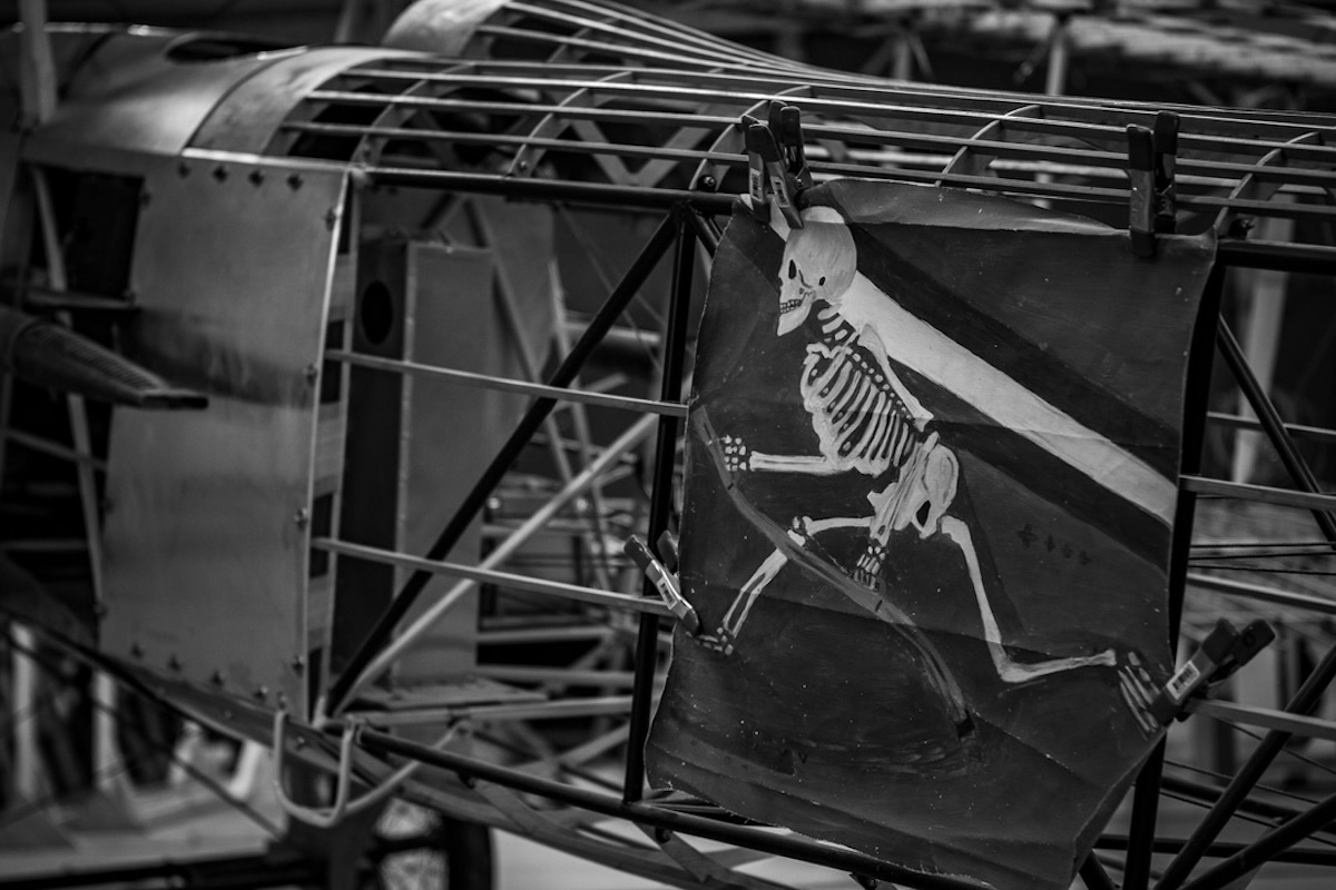 A black and white photo of the uncovered fuselage framework of a replica SPAD S.XIII. The image is dark. Clipped to one of the upper longerons is a piece of fabric with a 'grim reaper' graphic: a skeleton with one of its hands. (📸 James Woodard | Full Disc Aviation)