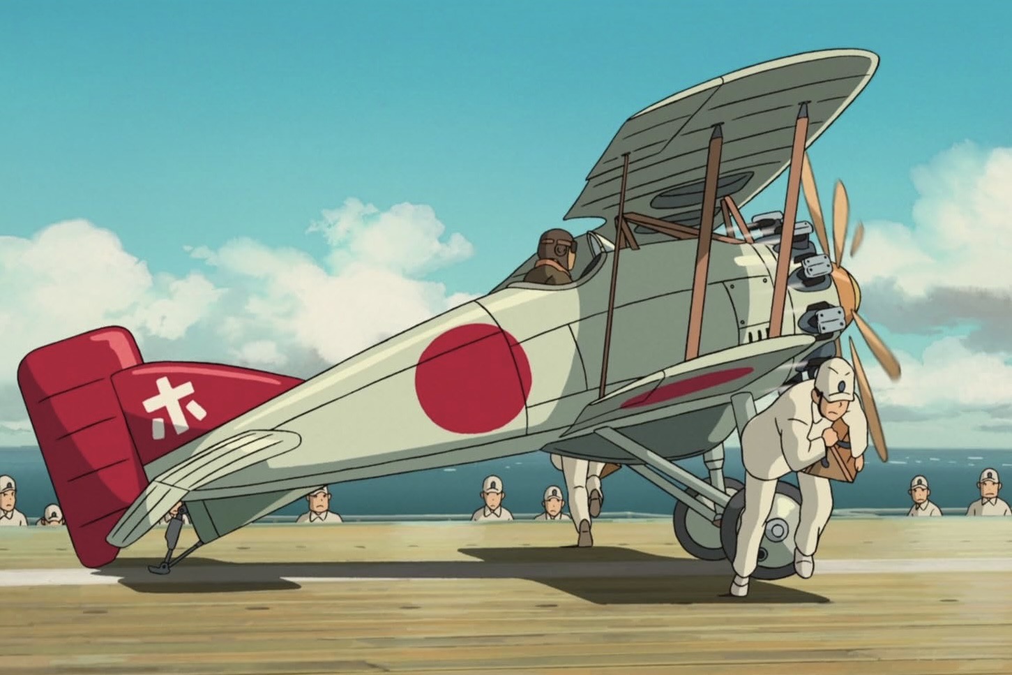A screenshot from the film 'The Wind Rises' wherein a biplane is shown just starting its take-off run from the deck of an aircraft carrier. The plane is moving from left to right in the frame. A crew member dressed in white is shown removing the wheel chocks from the aircraft. The aircraft is primarily white, with a red tail. A Japanese 'rising sun' roundel is painted on the fuselage. Other crew members are shown in the background watching the progress of the plane. The sky in the background is blue dotted with cumulus white clouds.  Original caption from source: « A Nakajima A1N1 is shown just before take-off during carrier trials. » (📸 ©2013 Hayao Miyazaki/Studio Ghibli, NDHDMTK)