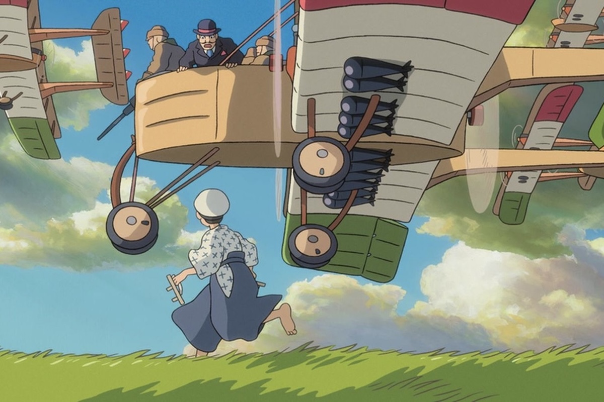 A screenshot from the film 'The Wind Rises' wherein a Caproni Ca.3 flies low over a green field from right to left in the frame. From the cockpit area, a man with a bowler hat and a moustache looks down at a boy running alongside the aircraft. The boy looks up at the plane. The boy is wearing traditional Japanese clothing, including traditional 'geta' footwear. Another Ca.3 is visible in the background. The sky is blue, dotted with puffy white clouds.  Original caption from source: « In a dream sequence, Count 'Gianni' Caproni, aboard his Caproni Ca.3, waves at the boyhood Jiro Horikoshi, who is running alongside on the ground. In particular, note the shoes in Jiro's hands. » (📸 ©2013 Hayao Miyazaki/Studio Ghibli, NDHDMTK)