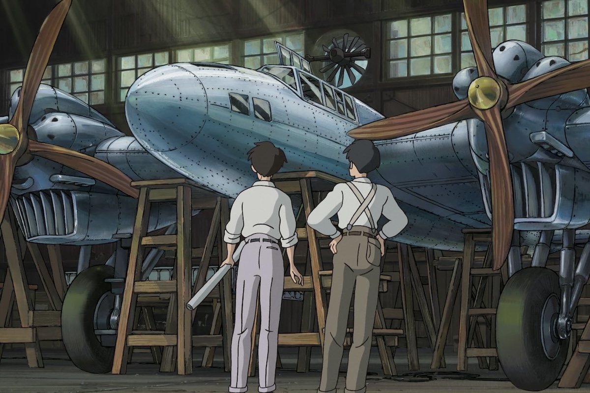 A screenshot from the film 'The Wind Rises' wherein two animated characters stand in front of a large, metal aircraft.  Original caption from source: « In a dimly lit factory, Jiro Horikoshi inspects a prototype of the Mitsubishi G1M1 long-range twin-engined attack bomber. » (📸 ©2013 Hayao Miyazaki/Studio Ghibli, NDHDMTK)
