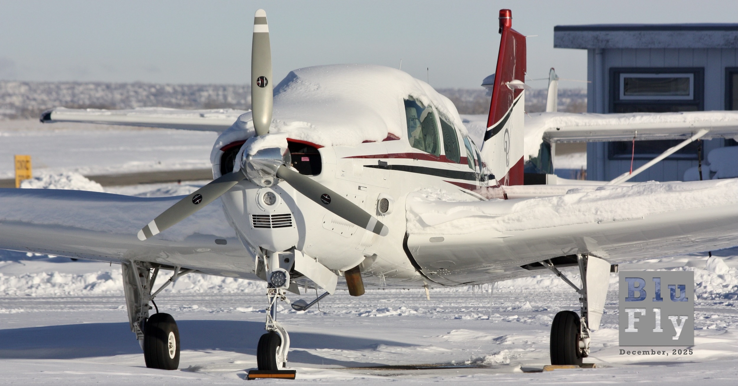 A Beech Debonair — that is, the straight-tail version of the Beech Bonanza — is pictured from the front and slightly to its left while parked at Springbank Airport near Calgary, Alberta, Canada. It is a tight crop focused on the fuselage, tail, and landing gear while clipping off the aircraft's wingtips. The upper surfaces of the aircraft are covered in a thin (ten centimetres?) layer of freshly fallen snow. The aircraft itself is white with red-and-blue accent stripes. The stylised 'B' from the Beech logo is visible on the vertical stabiliser. The aircraft has a three-bladed propeller. The ground around the aircraft is also covered in snow. In the background and to the right of the frame is a hangar and other aircraft. In the far background, what appears to be a housing development can be seen on the horizon. The sky above is a light, hazy blue but otherwise clear of cloud. Winter arrived early at Springbank Airport near Calgary, Alberta, Canada, as captured in November, 2024. (📸 ©2024 Terence C. Gannon, all rights reserved.)  Original caption from source: « Winter arrived early at Springbank Airport near Calgary, Alberta, Canada, as captured in November 2024. » (📸 ©2024 Terence C. Gannon. All rights reserved.)
