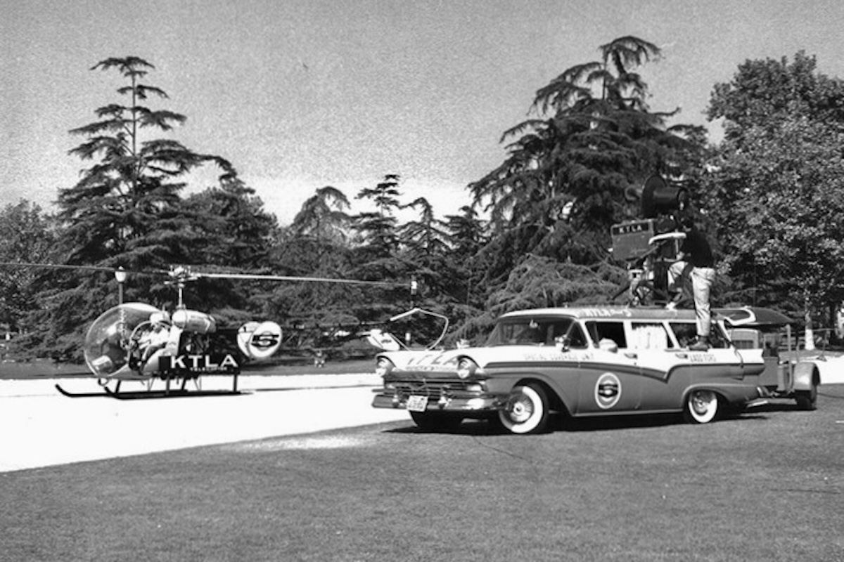 A black and white photo of a Bell 47-type helicopter parked on a helipad on the left side of the frame. The letters « KTLA » are painted on its side. On the right side of the frame, a station wagon is pictured. It also has the letters « KTLA » painted on the side. A television camera and cameraman are on the roof of the car with the camera pointing at the helicopter. A line of trees is visible in the background.  Original caption from source: « From 1958 via the 'Eyes of a Generation' website, the KTLA 'Telecopter' meets up with a KTLA-branded vehicle. » (📸 KTLA)