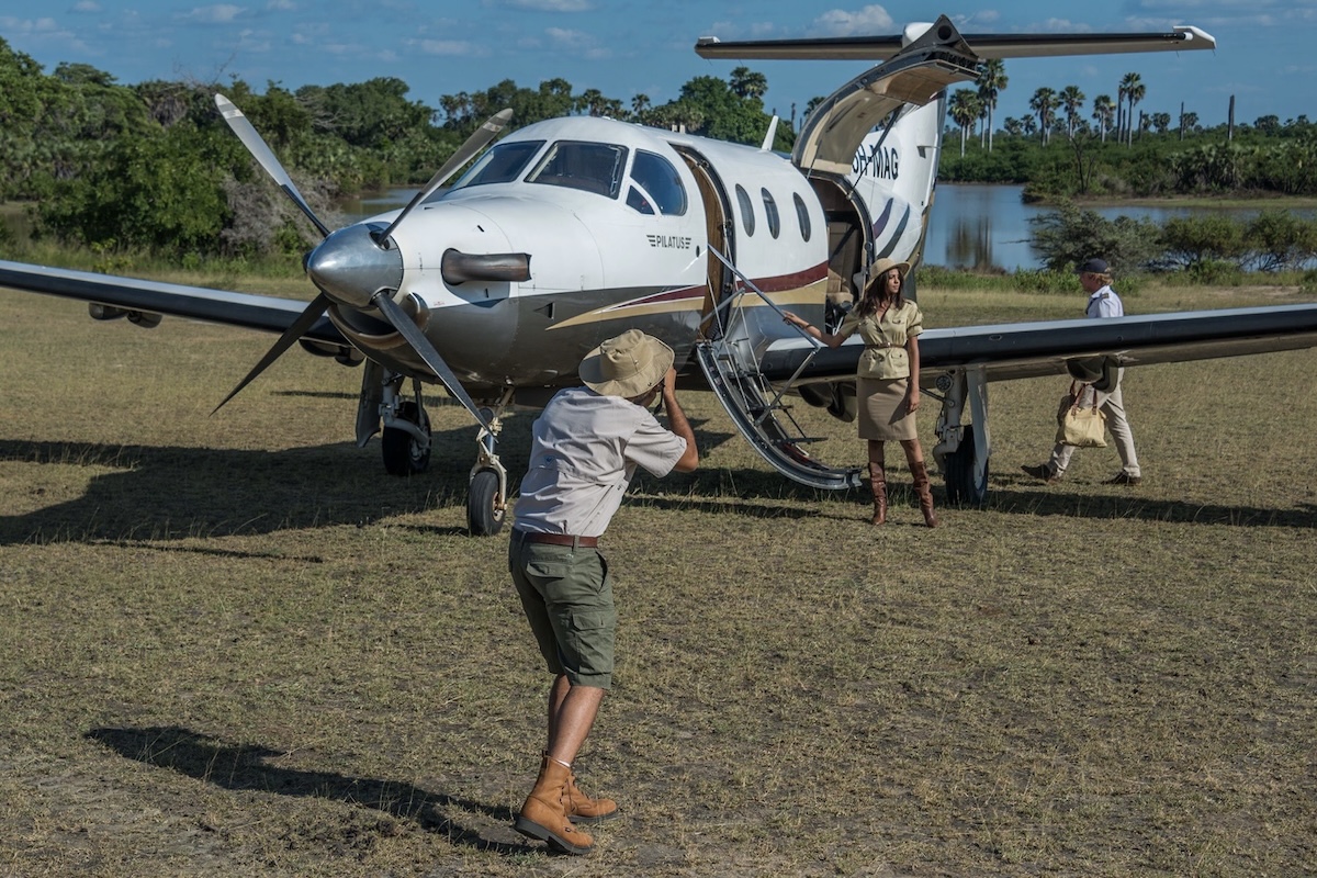 A man takes a photograph of a Pilatus PC-12 while a woman model stands at the stairs leading up to the aircraft. She is wearing 'safari' garb. A man in the background is loading luggage into the large open cargo door aft of the aircraft's wing. The aircraft, registration « 5H-MAG », is oriented toward and slightly to the left of the camera and is parked on a grass ramp. A blue lake is visible in the background. Palm trees are visible in the line of trees beyond the lake. The sky above is blue with white puffy clouds. (📸 ©2017 Jon Davison | Eye in the Sky Productions)