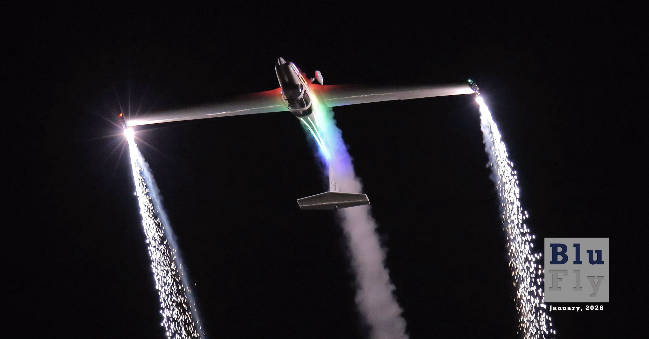 An all-white motorglider, separately identified as a Grob G109B, ascends through the top of a loop during a night display flight. Sparks stream from each wingtip and white smoke trails from behind the fuselage. The aircraft is travelling directly upwards and slightly 'over the top' on its way to inverted flight and presumably through to complete the loop. The background is pitch black making for a dramatic backdrop for the aircraft. (📸 ©2016 Richard Davies | SupaSmokey)