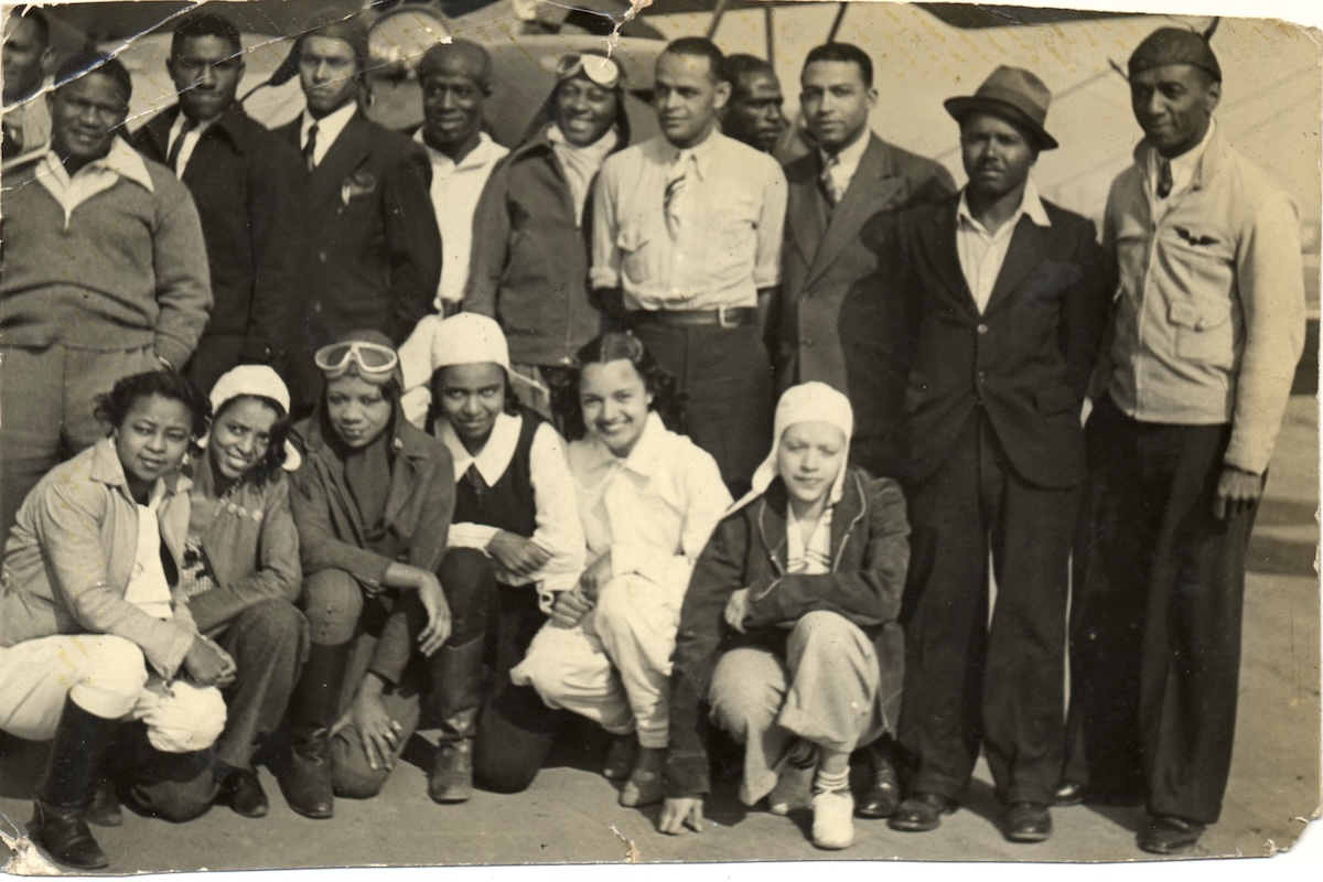A sepia-toned, black and white photo of a group of African-American aviators posing in two rows for the camera. Men are in the back row, and the women are crouching down in the front row. A number of them are wearing aviator garb such as cloth or leather flying helmets and goggles. Just visible in the background is a biplane. The image is cracked and broken down in a number of places, which attests to its age and condition. Original caption from source: « William J. Powell (far right), a successful owner of several automobile service stations in Chicago, moved to Los Angeles to learn to fly. By the early 1930s, Powell had organized the Bessie Coleman Aero Club to promote aviation awareness in the Black community. Both men and women were welcome to apply. Powell became a talented visionary and promoter of Black involvement in aviation. » (📸 National Air & Space Museum)