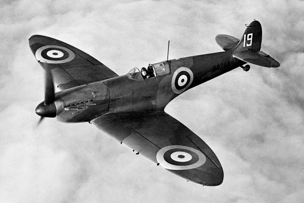 A camouflaged Spitfire low-wing monoplane fighter in flight captured from above and ahead of the aircraft. A solid, cumulus cloud deck appears in the background.