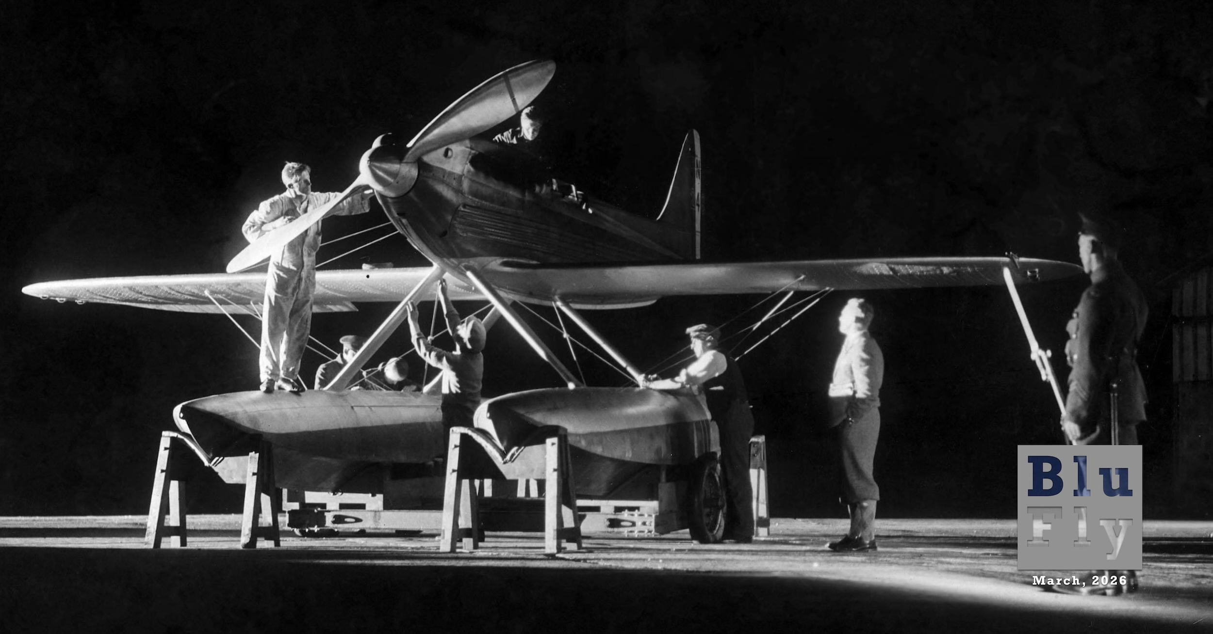 A low-wing, monoplane aircraft with two pontoons in a hangar at night. It is lit with spotlights, and a group of men are performing maintenance. (📸 Mirrorpix | Alamy under license.)