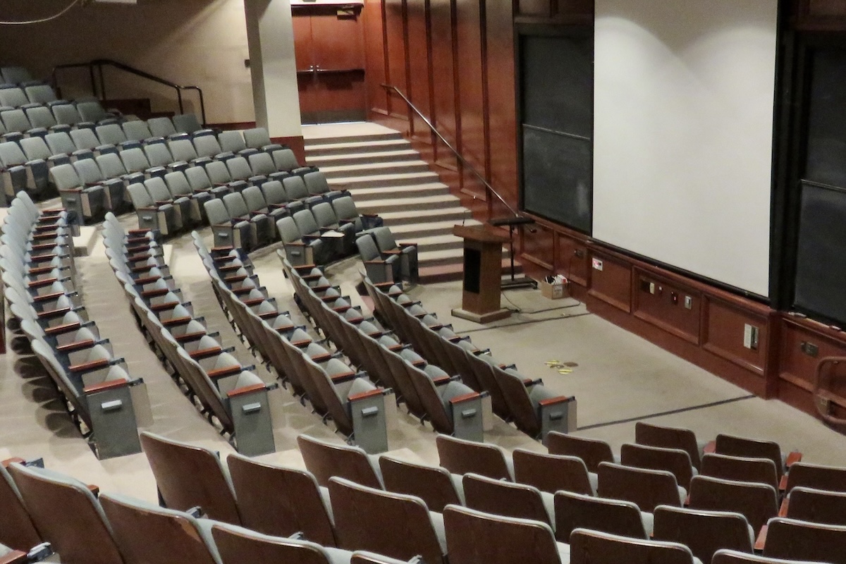 An empty university auditorium. The lectern is in the middle, and blue seats slope upwards to the left. A large screen is on the right.