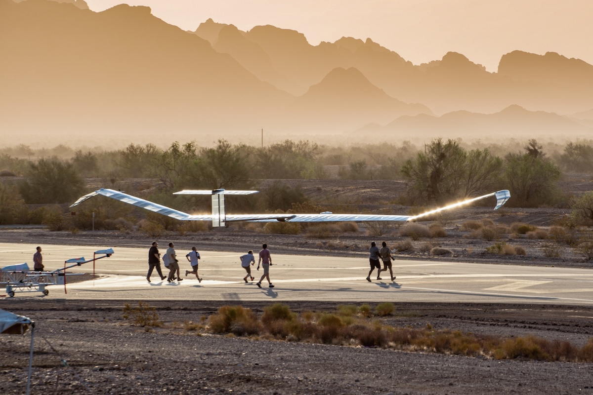 Ground handlers run below an Airbus/AALTO Zephyr flying a few metres above a runway in a desert landscape. It is either sunrise or sunset.