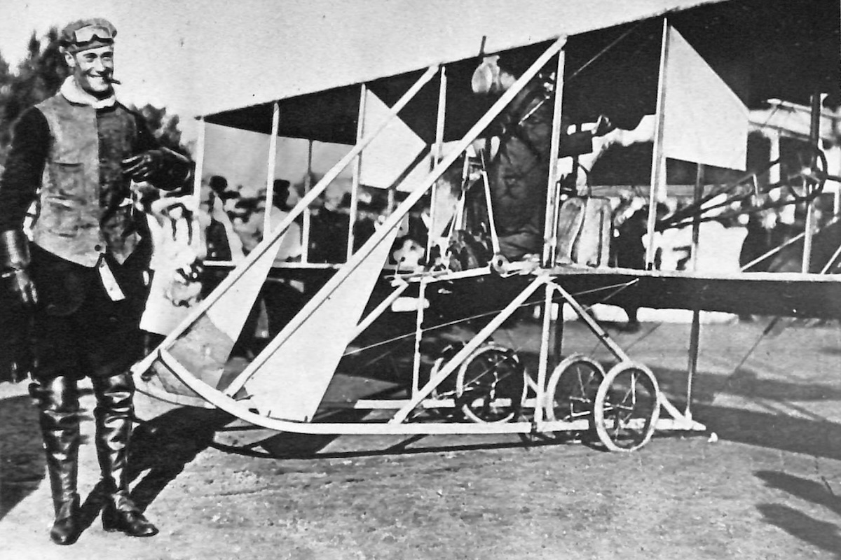 A man in 1910s-era aviation clothing in front of a delicate-looking biplane. Another man is engaged in some sort of activity in the pilot's seat.