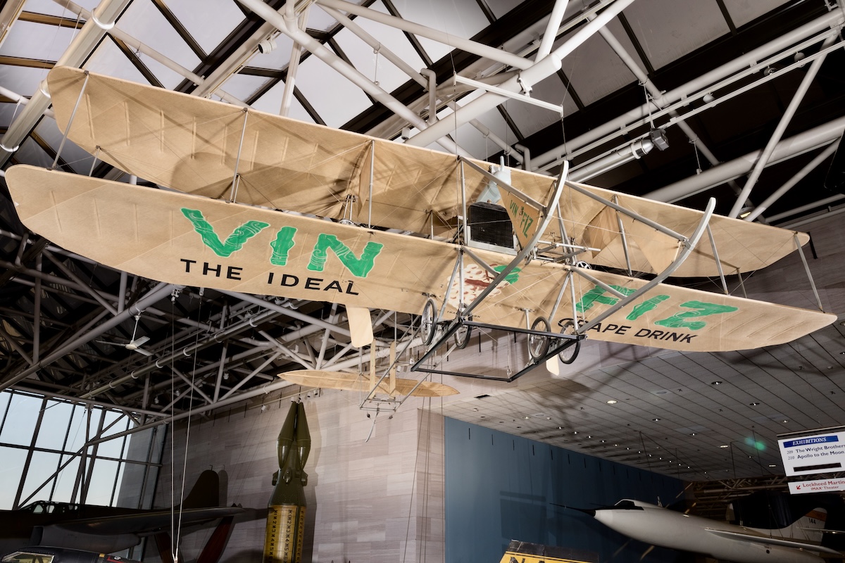 A delicate-looking biplane suspended from a framework ceiling with « VIN FIZ » painted in large, green letters on the underside of the lower wing.
