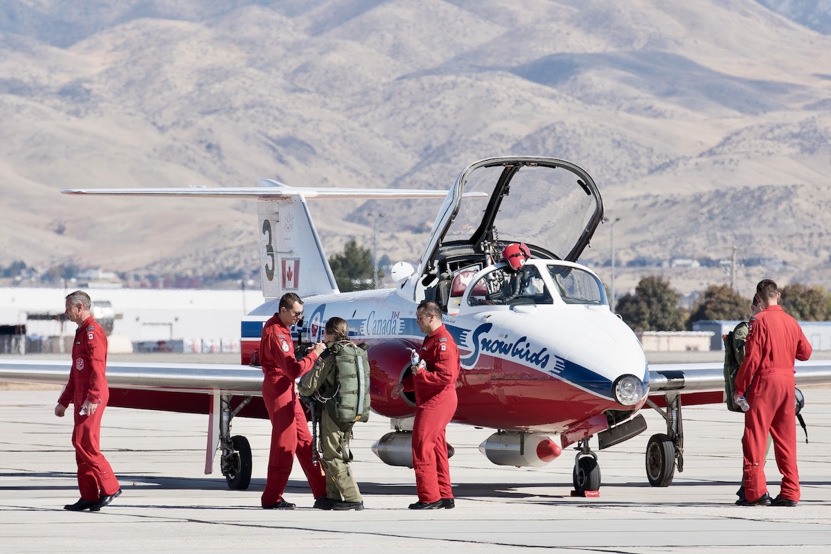 A member of the RCAF Snowbirds is parked on a runway in a desert landscape. Members of its crew, in red jumpsuits, surround it.