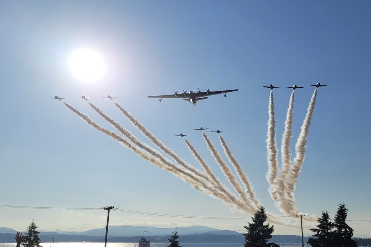 A Martin Mars flying boat flies toward the camera position under clear, sunny skies in formation with a nine-plane formation of the RCAF Snowbirds.