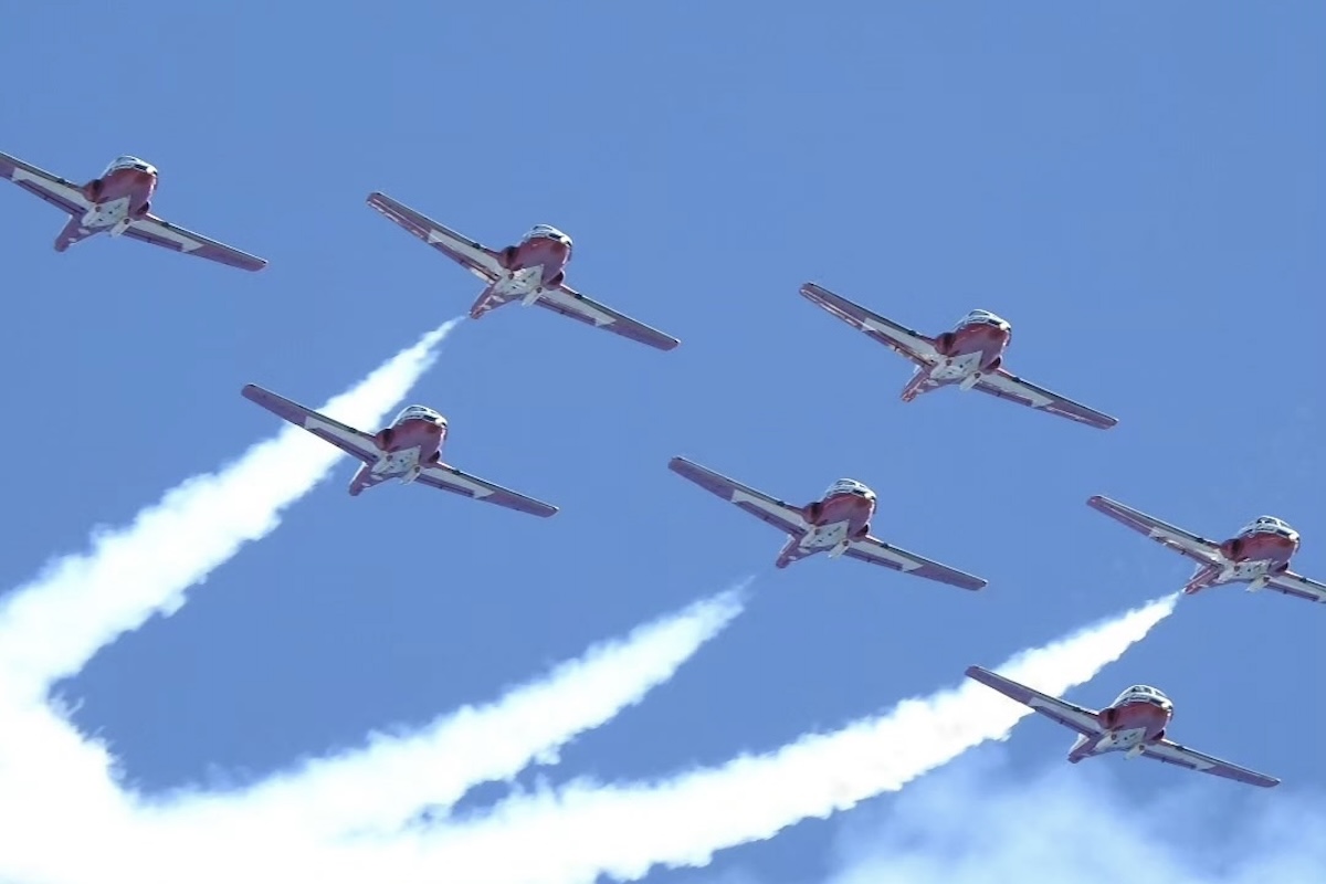 A tight, seven-plane formation of the RCAF Snowbirds, flying toward the camera, starts a barrel roll in a blue sky while trailing white smoke.