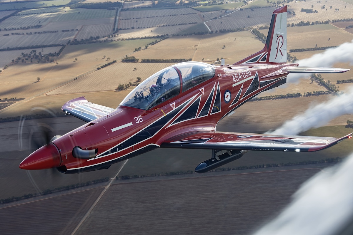 An air-to-air photo of a Royal Australian Air Force PC-21 in the livery of the Roulettes demonstration team, trailing white smoke. The background is farmland.