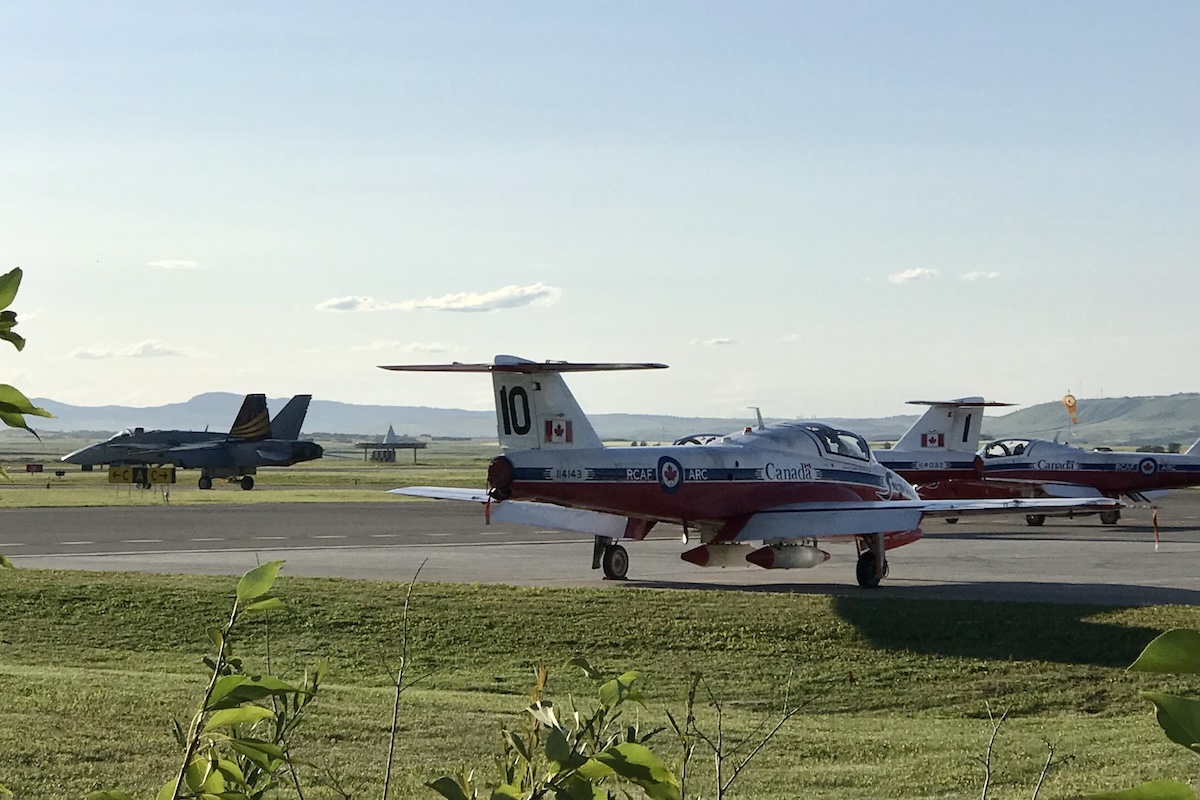 Three members of the RCAF Snowbirds park on the ramp at a country airport. A CF-18 taxies by them, in the distance.
