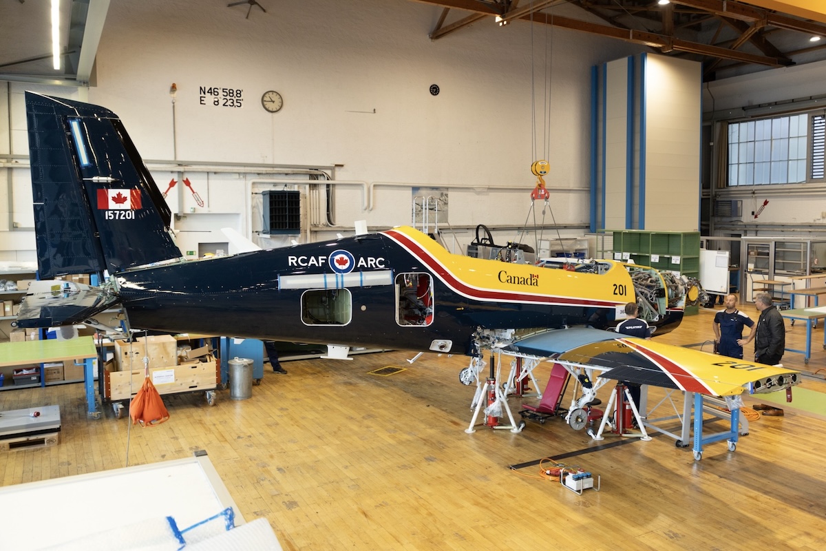 A Pilatus CT-157 Siskin II is pictured under construction in a well-lit, spotlessly clean manufacturing facility. To its right, two workers are in a discussion.