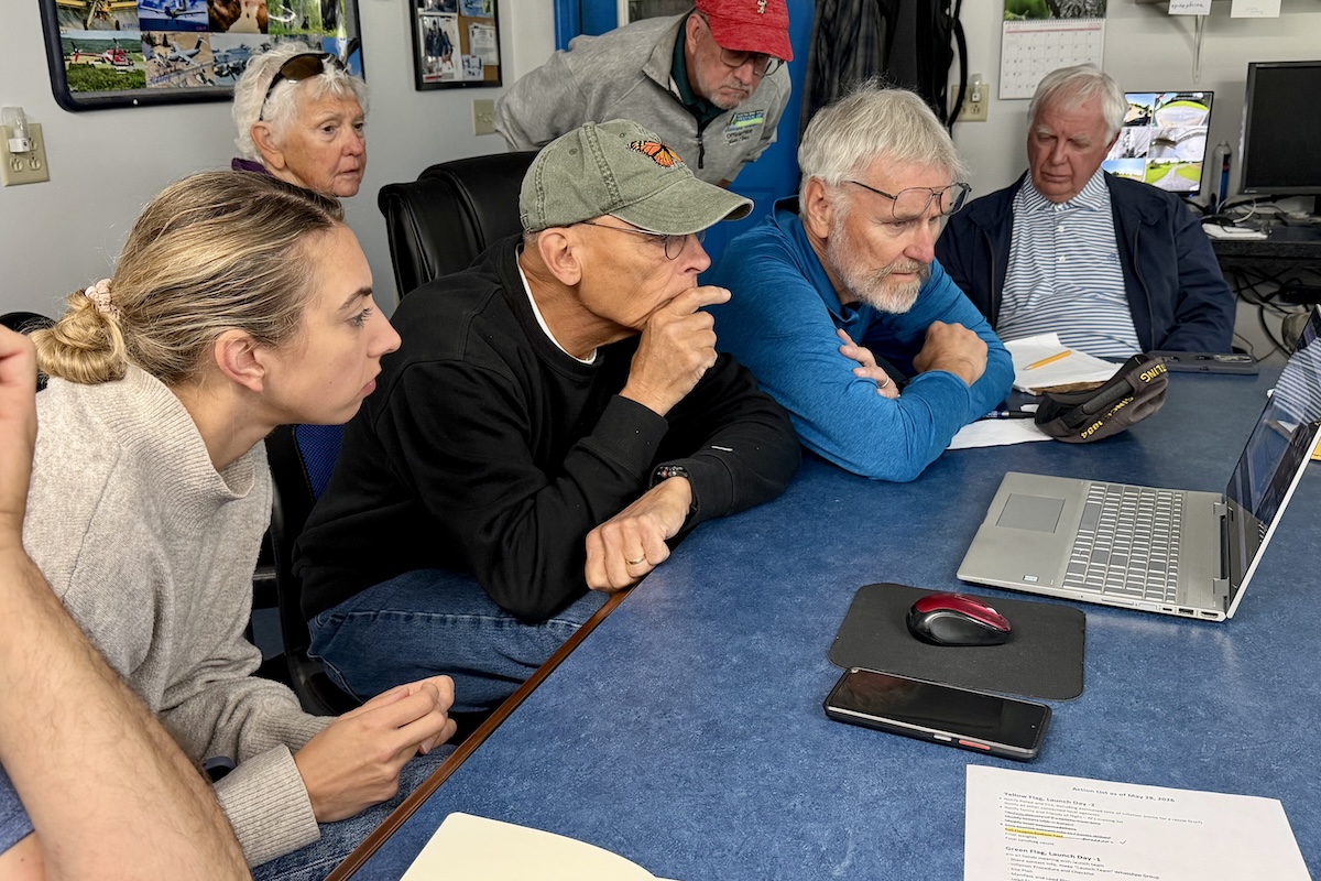 A group of people sit or stand behind a table with a laptop on it, and they are all looking at what's on the screen.