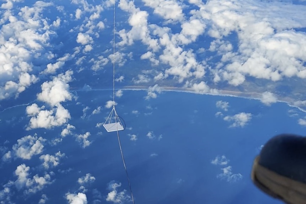 The view from a balloon gondola on a brightly lit day. The ocean and a coastline appear, and the ground is partially obscured by clouds.