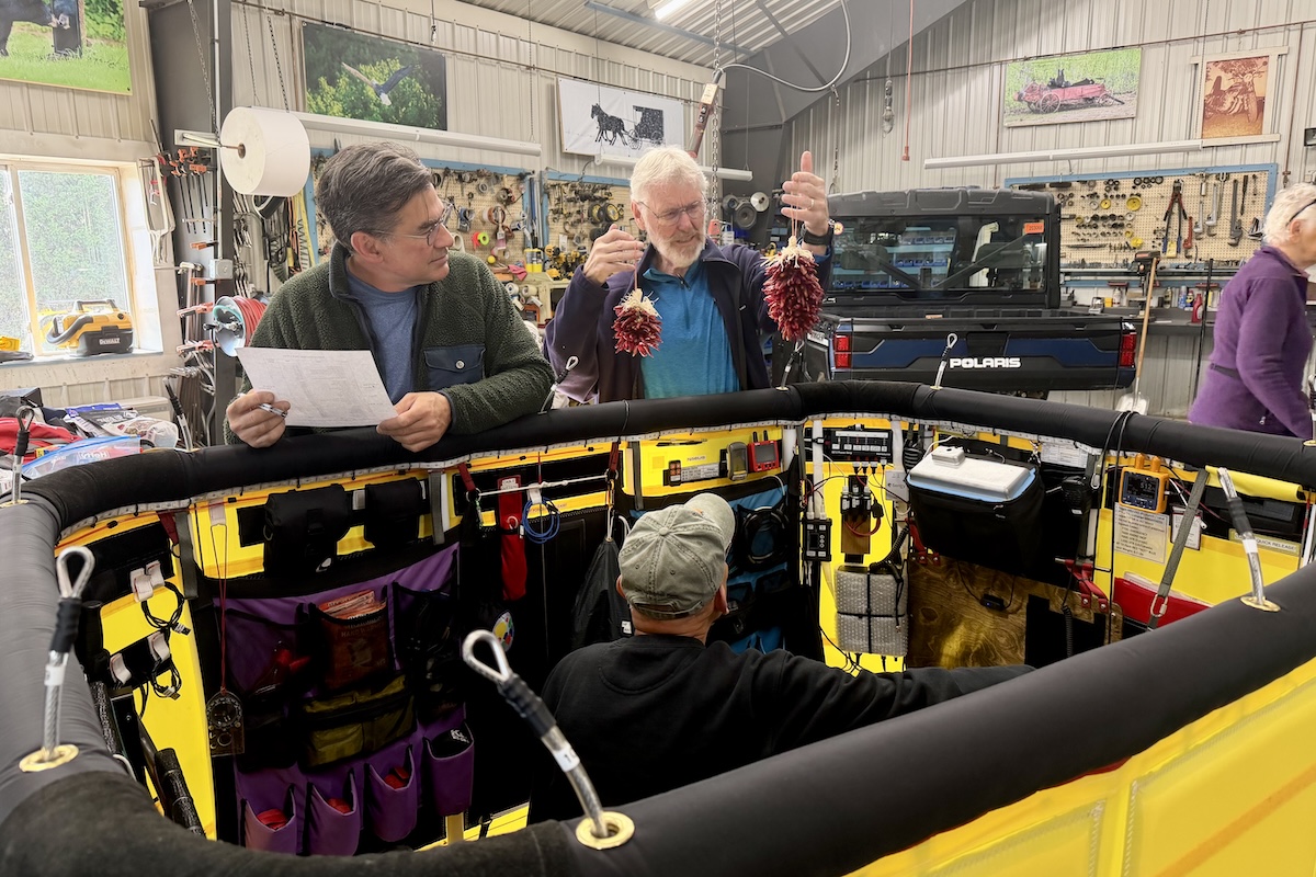 Three people work in and around a balloon gondola inside a workshop. One is holding up a chile ristra so as to weigh it.