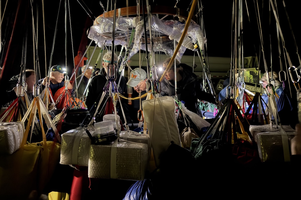 Three people stand inside a balloon gondola, and many more stand outside, all working intently under floodlights. A number of ropes extend upwards.