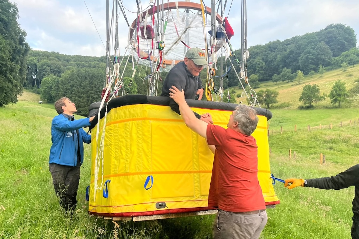 A man in a balloon gondola that has just landed in a field. He shakes hands with another man who is standing on the ground.
