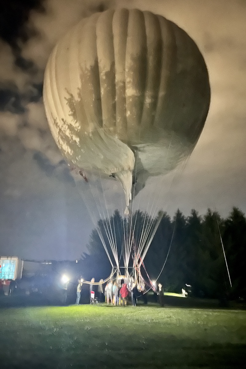 A gas balloon about to rise from a green field. Floodlights illuminate it. The shadows of trees appear on the side of the gas cell.
